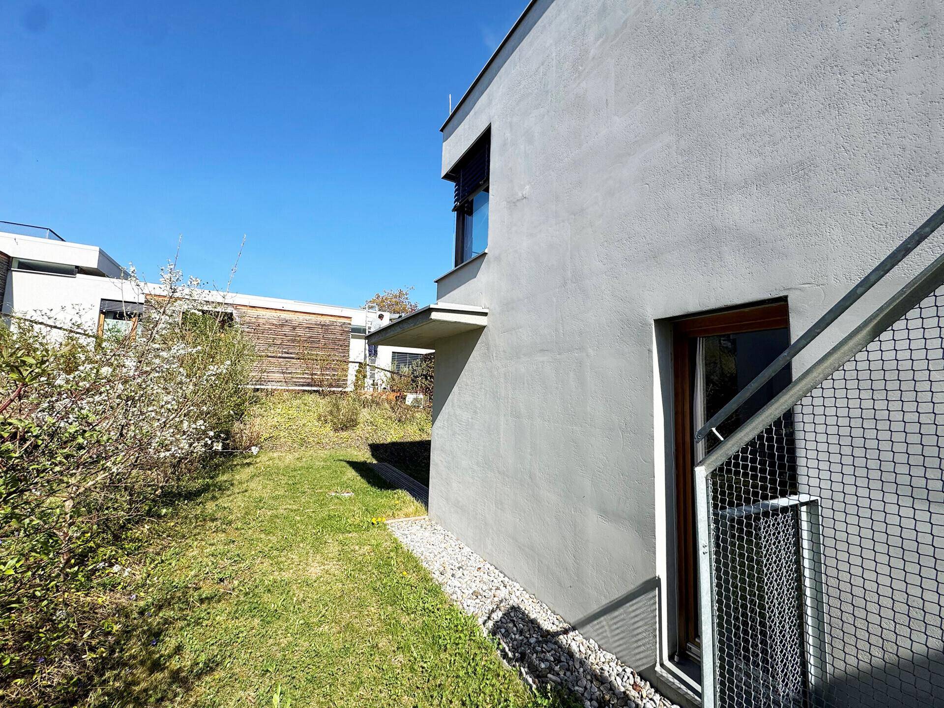 Seitenansicht des Gebäudes mit grünem Garten und Kiesstreifen unter blauem Himmel.