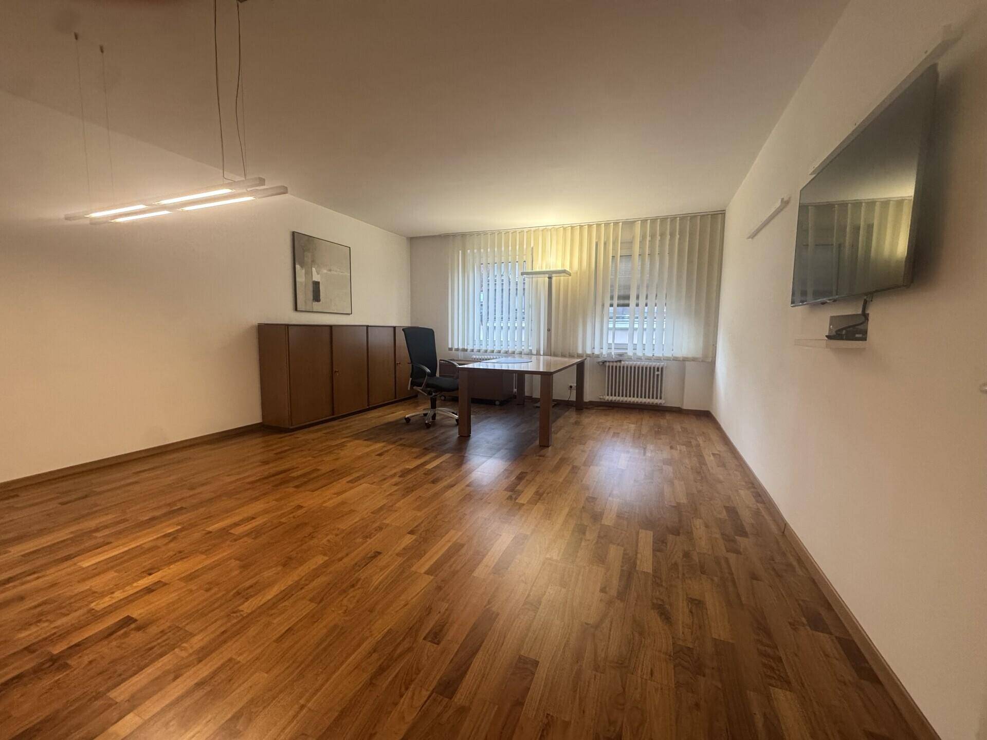 Geräumiges Büro mit Holzboden, großen Fenstern und moderner Beleuchtung, ausgestattet mit Schreibtisch und Schränken.