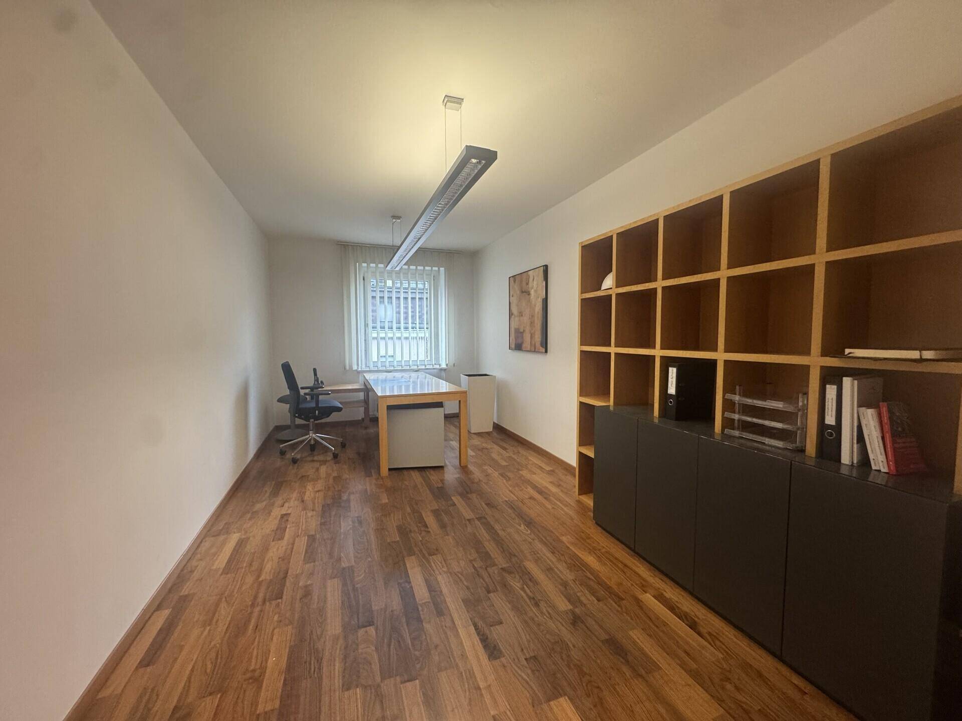 Büro mit Holzboden, Schreibtisch und einem großen, offenen Regal für Bücher und Akten, beleuchtet durch eine moderne Lampe.