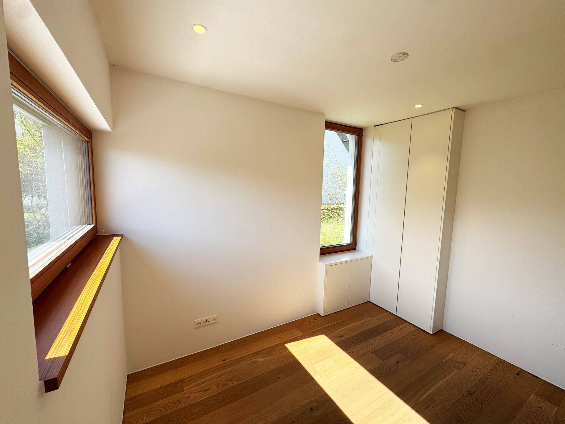 Helles Schlafzimmer mit Holzboden und zwei Fenstern, die viel Tageslicht hereinlassen.