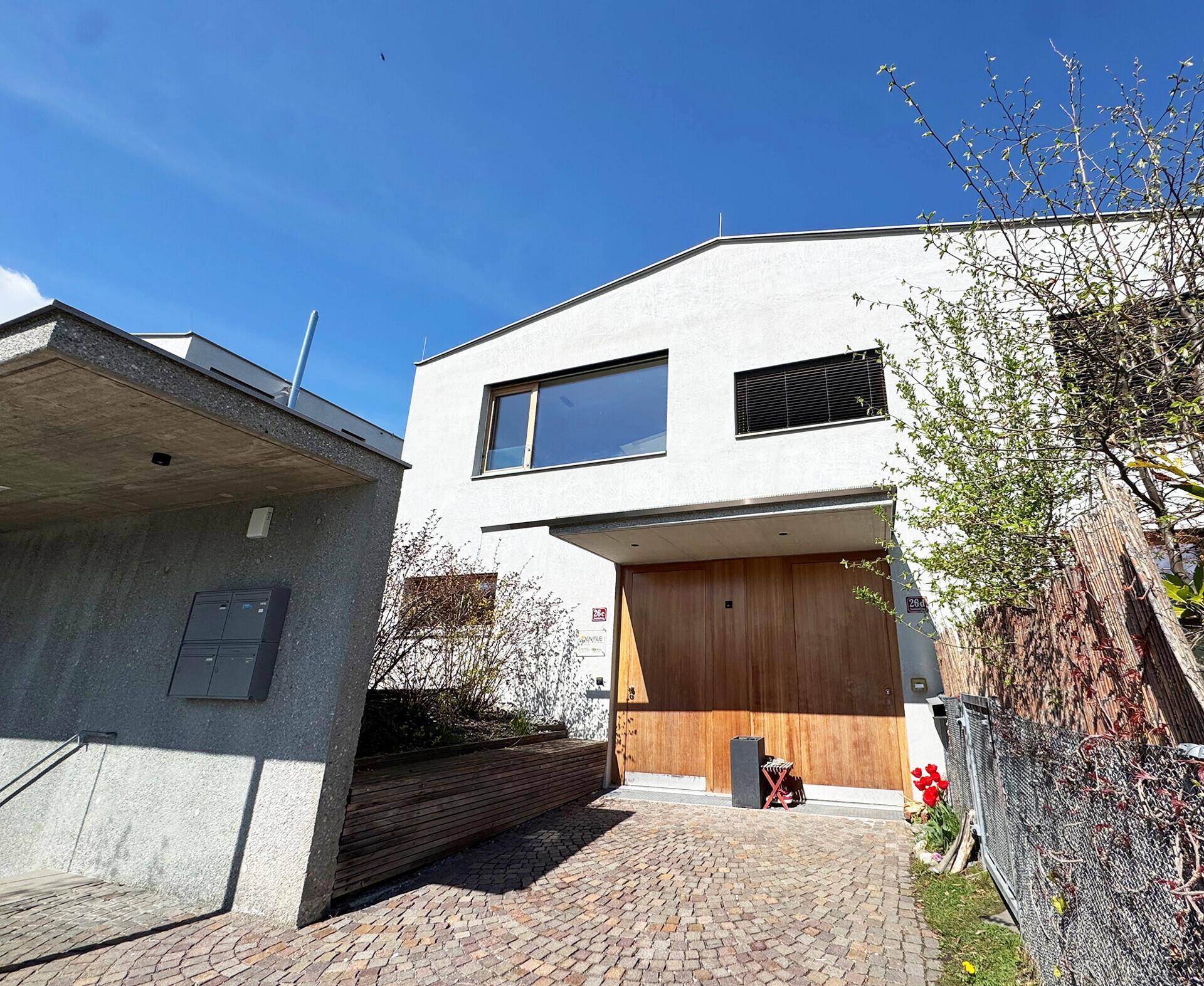 Moderne Hausfassade mit Holztor und gepflasterter Auffahrt unter strahlend blauem Himmel.