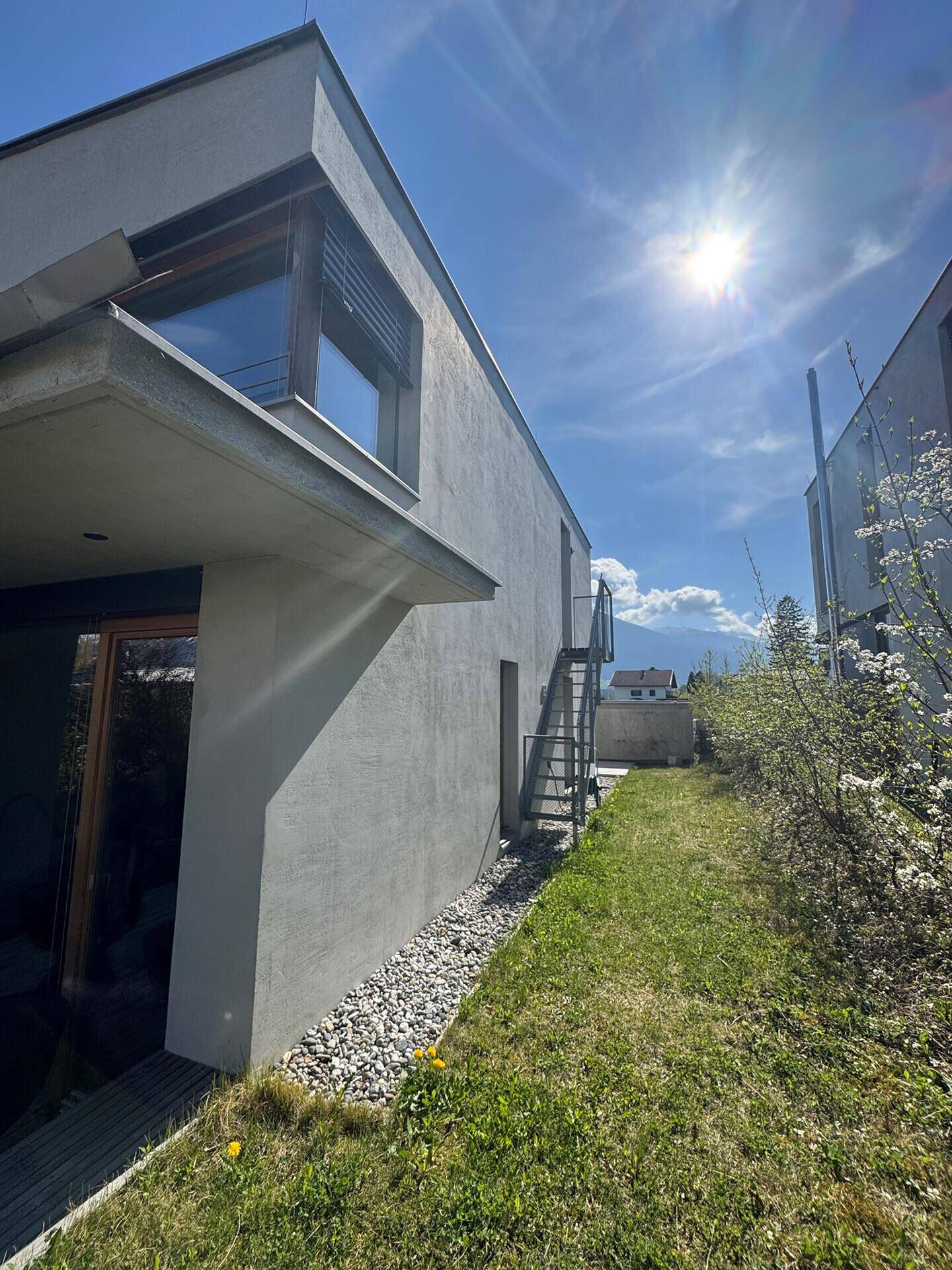 Seitenansicht des Hauses mit sonnigem Gartenbereich und Außentreppe unter blauem Himmel.
