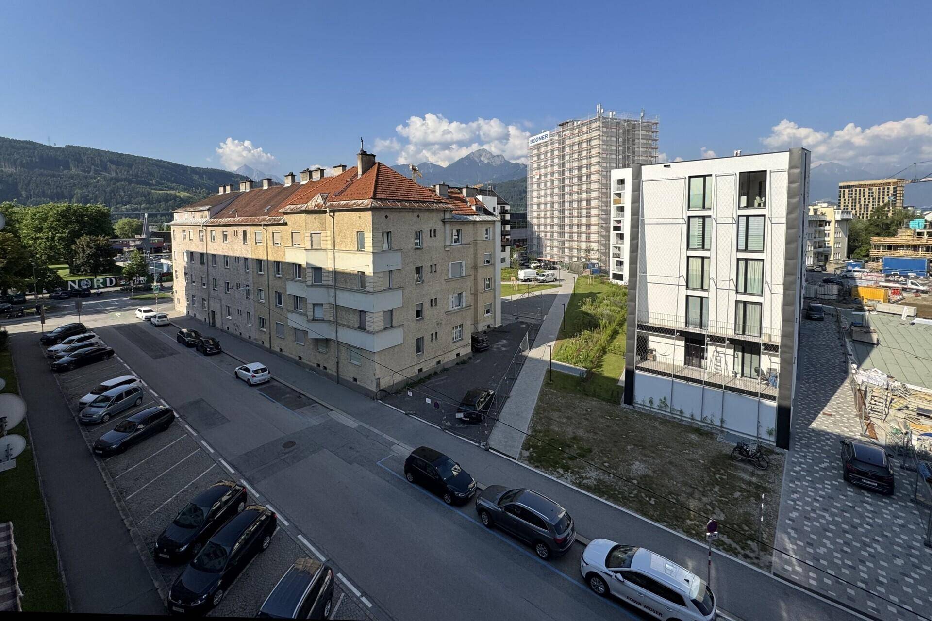Stadtansicht mit Gebäuden, geparkten Autos auf der Straße und Bergen am Horizont unter blauem Himmel.