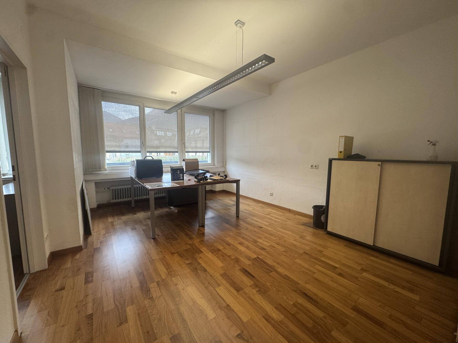 Büro mit Holzboden, großem Fenster und Schreibtisch, ergänzt durch einen Aktenschrank und eine moderne Deckenleuchte.