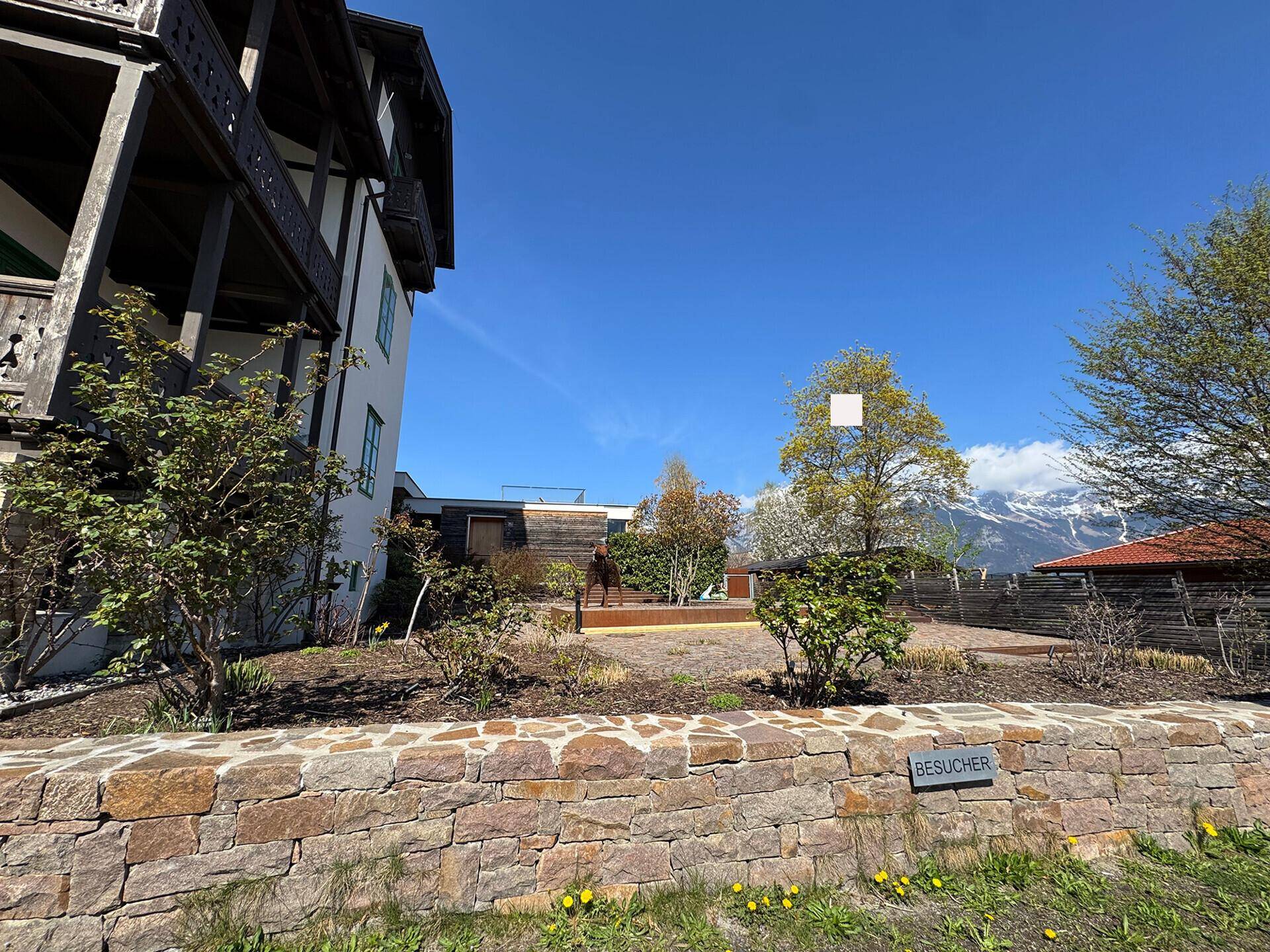 Gepflegter Gartenbereich mit Natursteinmauer und einem traditionellen Gebäude im Hintergrund.