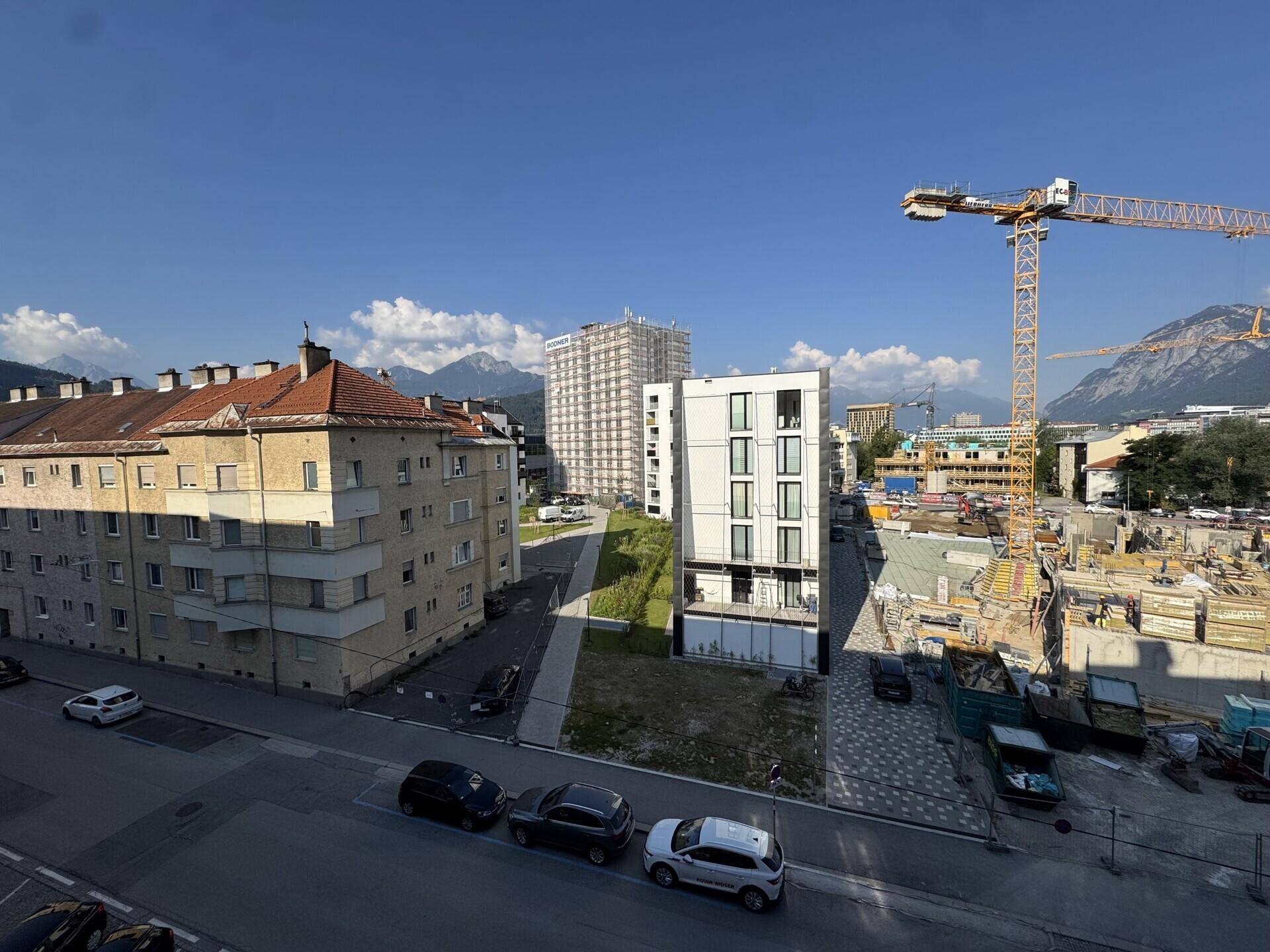 Panoramablick auf die Stadt mit Gebäuden, Baustelle und Bergen im Hintergrund an einem sonnigen Tag.