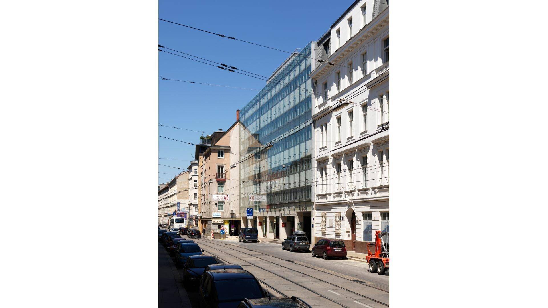 Weitläufige Straßenansicht mit dem modernen Glasgebäude und angrenzenden traditionellen Bauten unter blauem Himmel.