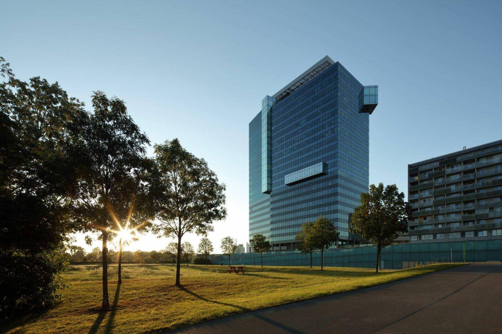 Modernes Hochhaus mit Glasfassade, umgeben von Grünflächen und Bäumen bei Sonnenuntergang.