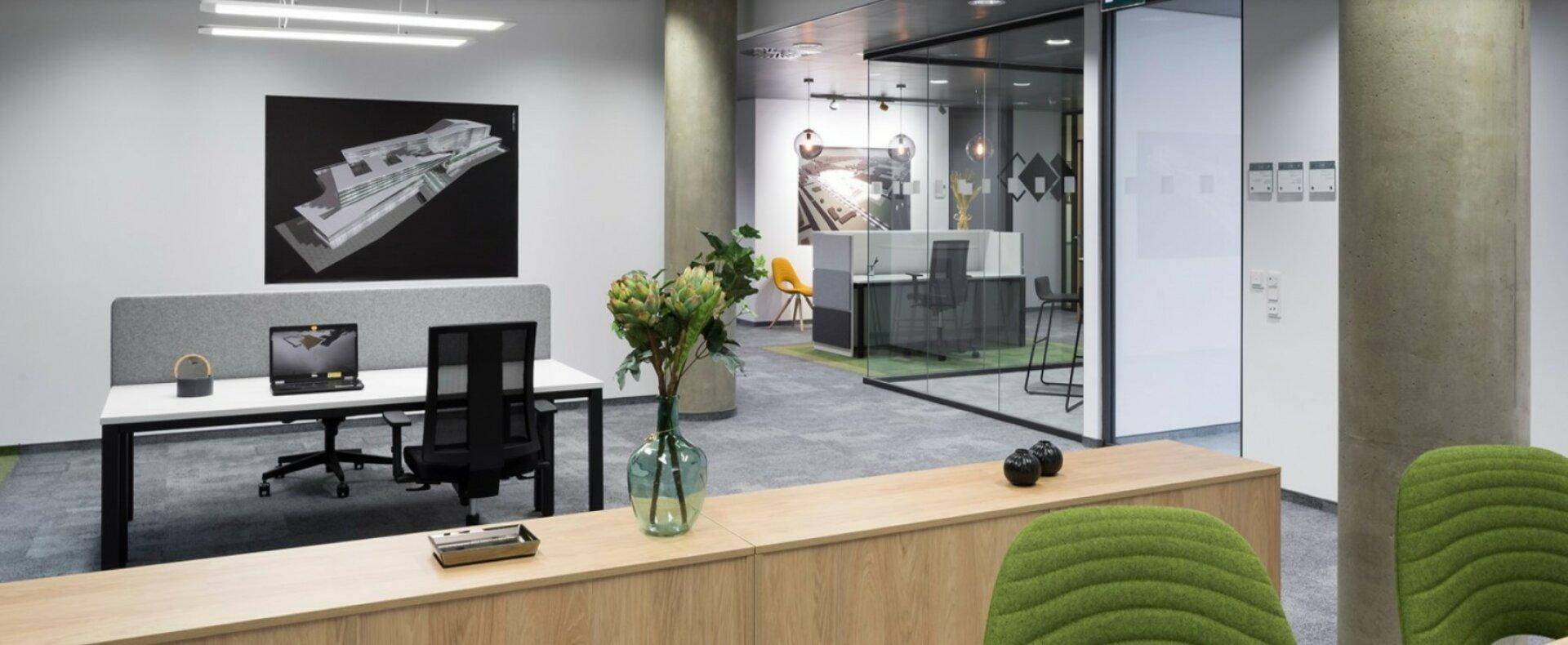 Moderner Büroraum mit Schreibtischen, grünen Stühlen und Glaswänden, die weitere Bürobereiche abtrennen.