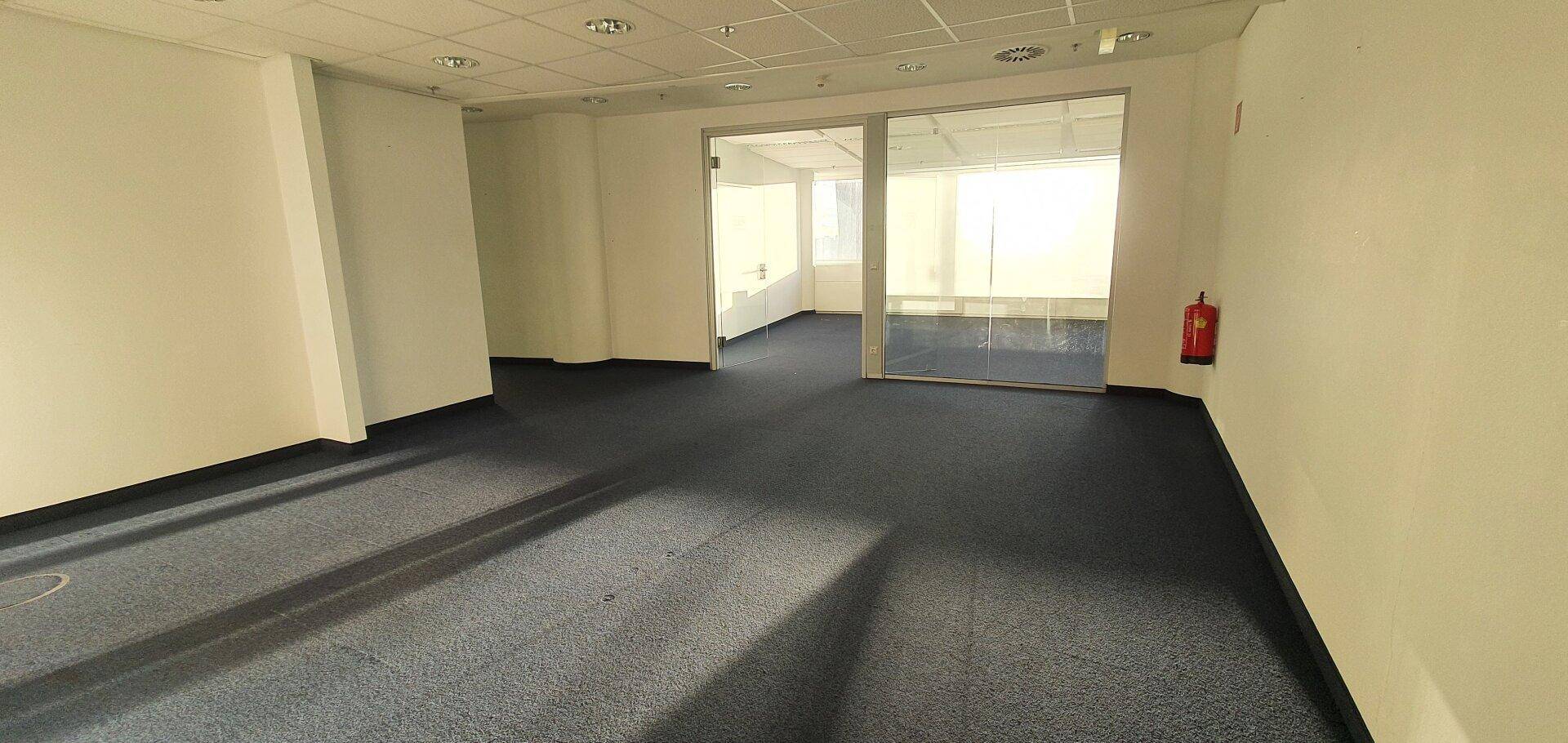 Heller Büroraum mit Teppichboden und einer Glastür, die zu einem weiteren Bereich führt.