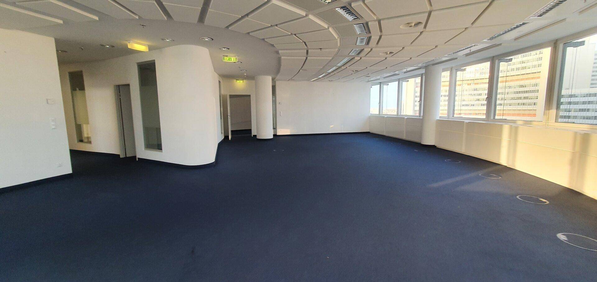 Großer, leerer Bürobereich mit Teppichboden und Zugang zu weiteren Räumen im Hintergrund.