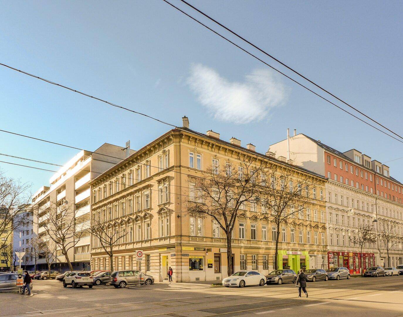 Historisches Eckgebäude an einer städtischen Kreuzung mit Geschäften im Erdgeschoss und parkenden Autos unter blauem Himmel.