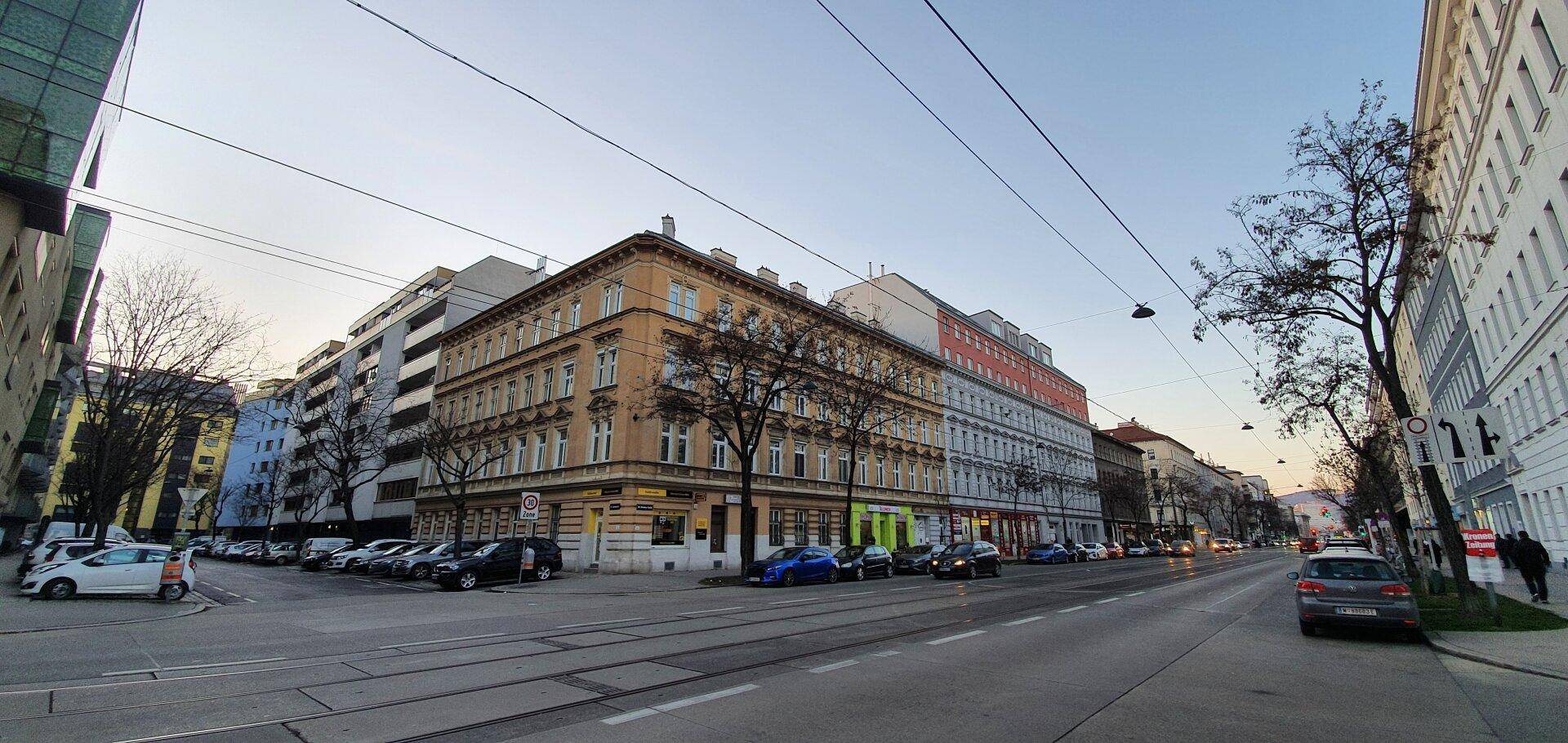 Weitwinkelaufnahme des historischen Gebäudes an einer belebten Stadtstraße mit Straßenbahnschienen und parkenden Autos.