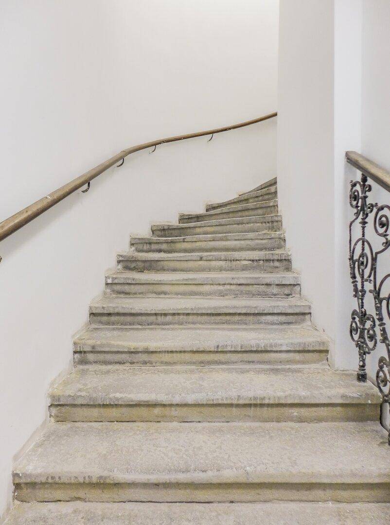 Geschwungene Steintreppe mit schlichtem Holzgeländer und schmiedeeisernen Details im historischen Gebäude.