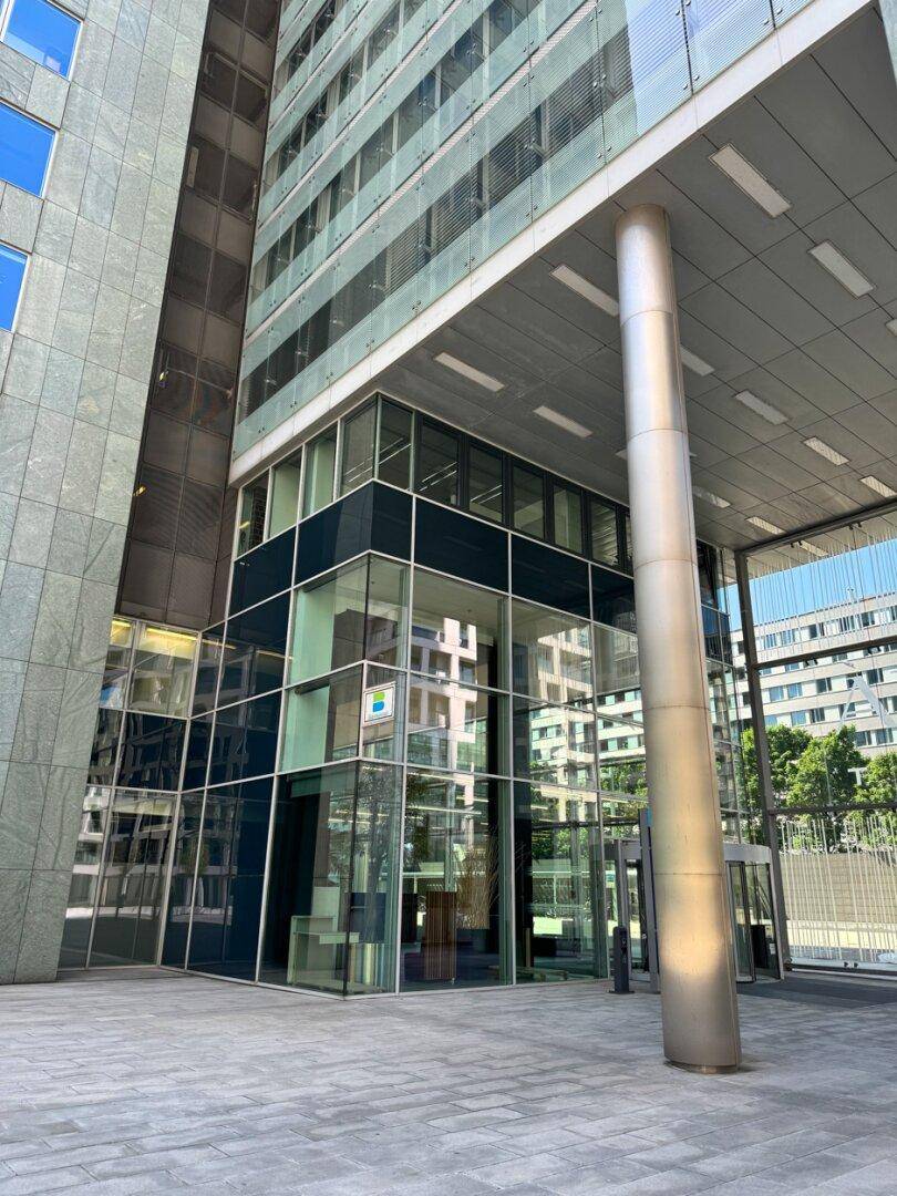 Eingangsbereich eines modernen Hochhauses mit großer Glasfront und markantem Stützpfeiler.