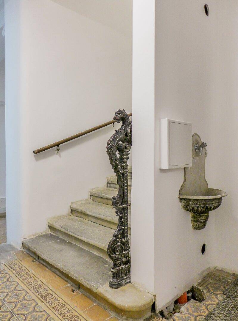 Historisches Treppenhaus mit Steinstufen, schmiedeeisernem Geländer und einem dekorativen Wandbrunnen.