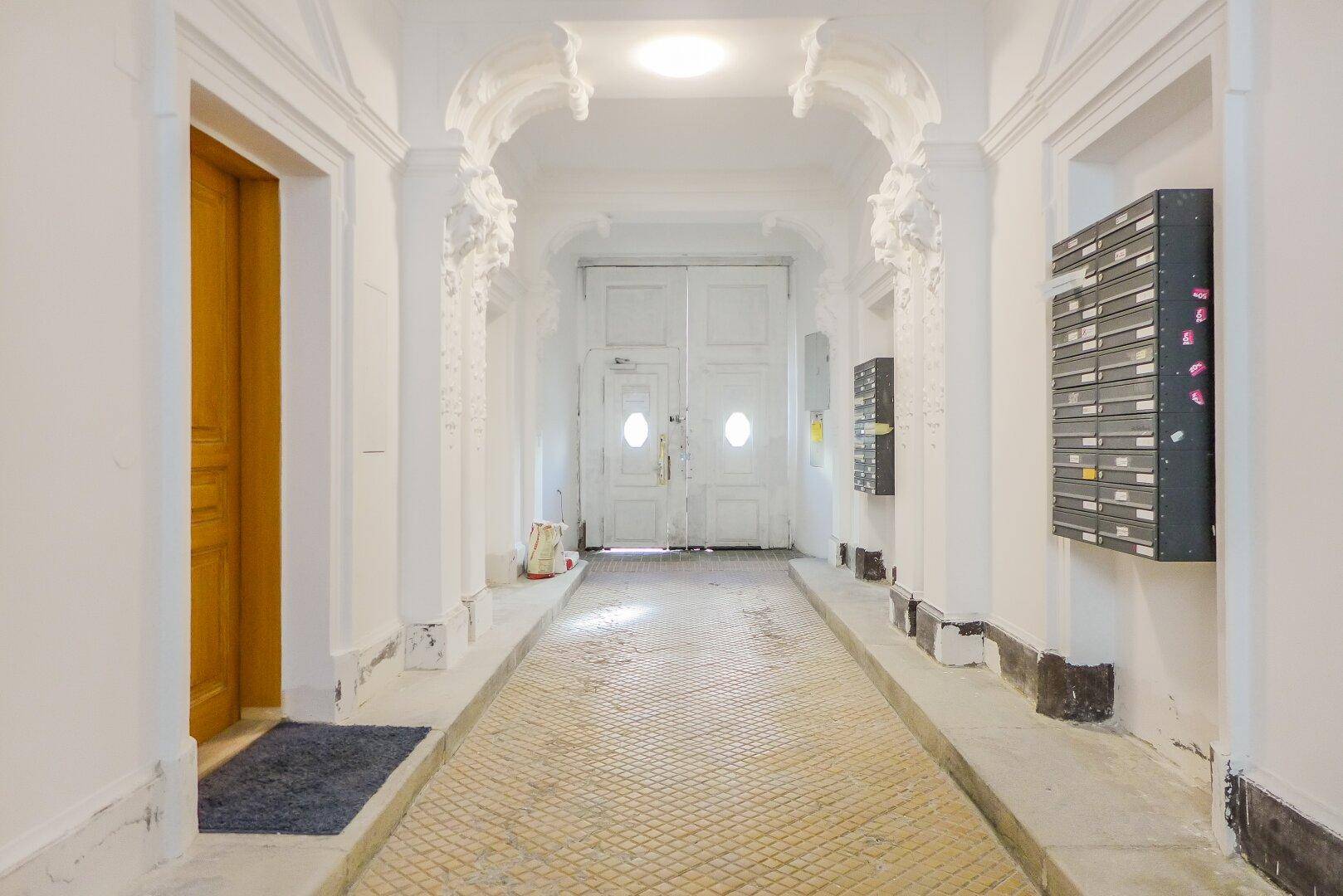 Langer Eingangsbereich mit historischen Stuckverzierungen, Fliesenboden und Briefkästen.