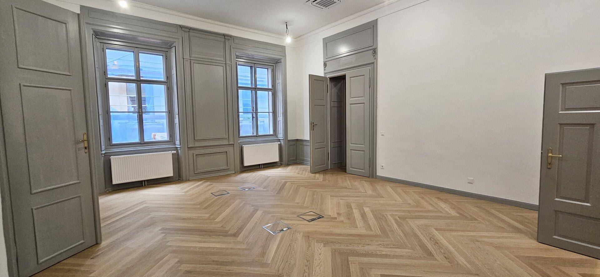 Heller Raum mit Fischgrätparkett, hohen Fenstern und dekorativen Wandpaneelen in Grau, bietet viel Tageslicht.