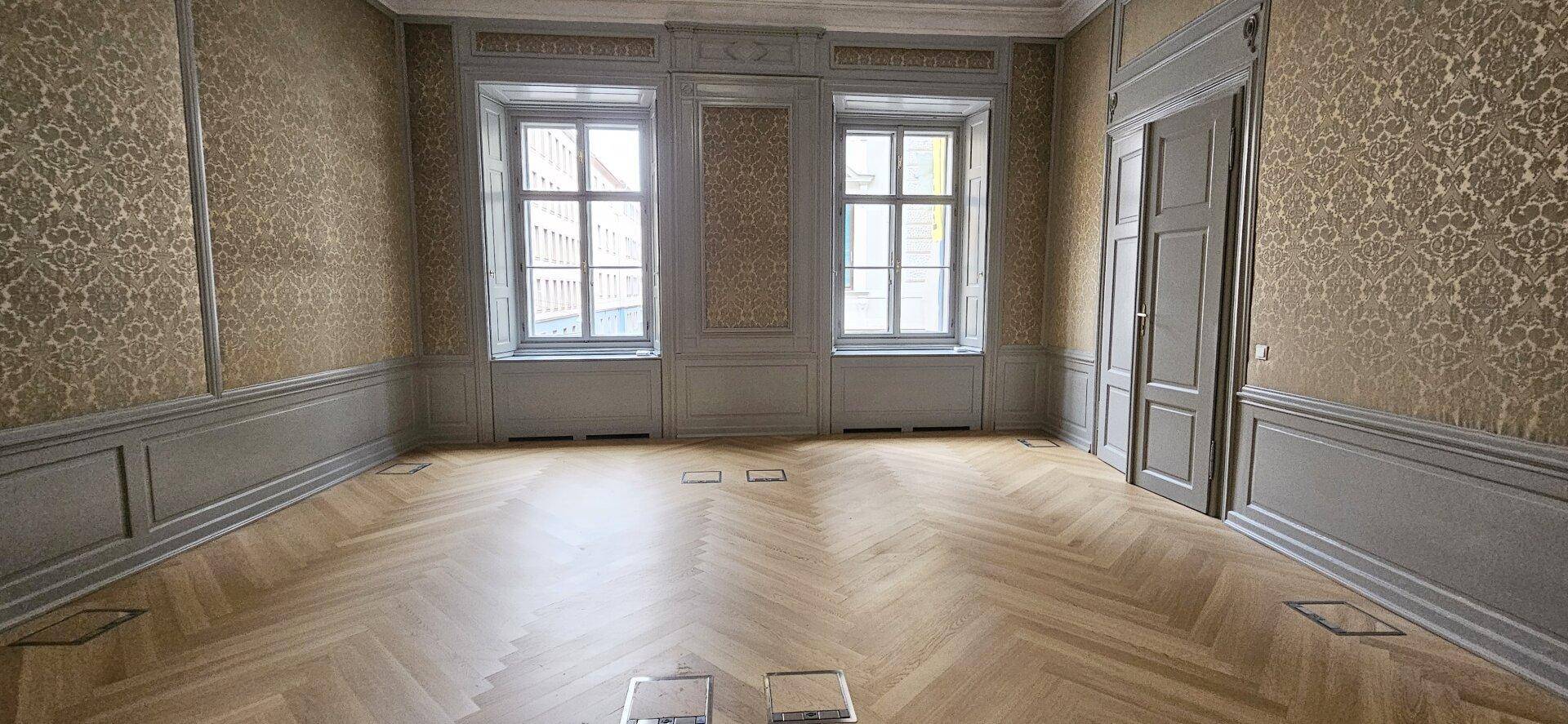Raum mit Fischgrätparkett, hohen Fenstern und gemusterten Tapeten sowie grauen Wandpaneelen, sehr elegant.