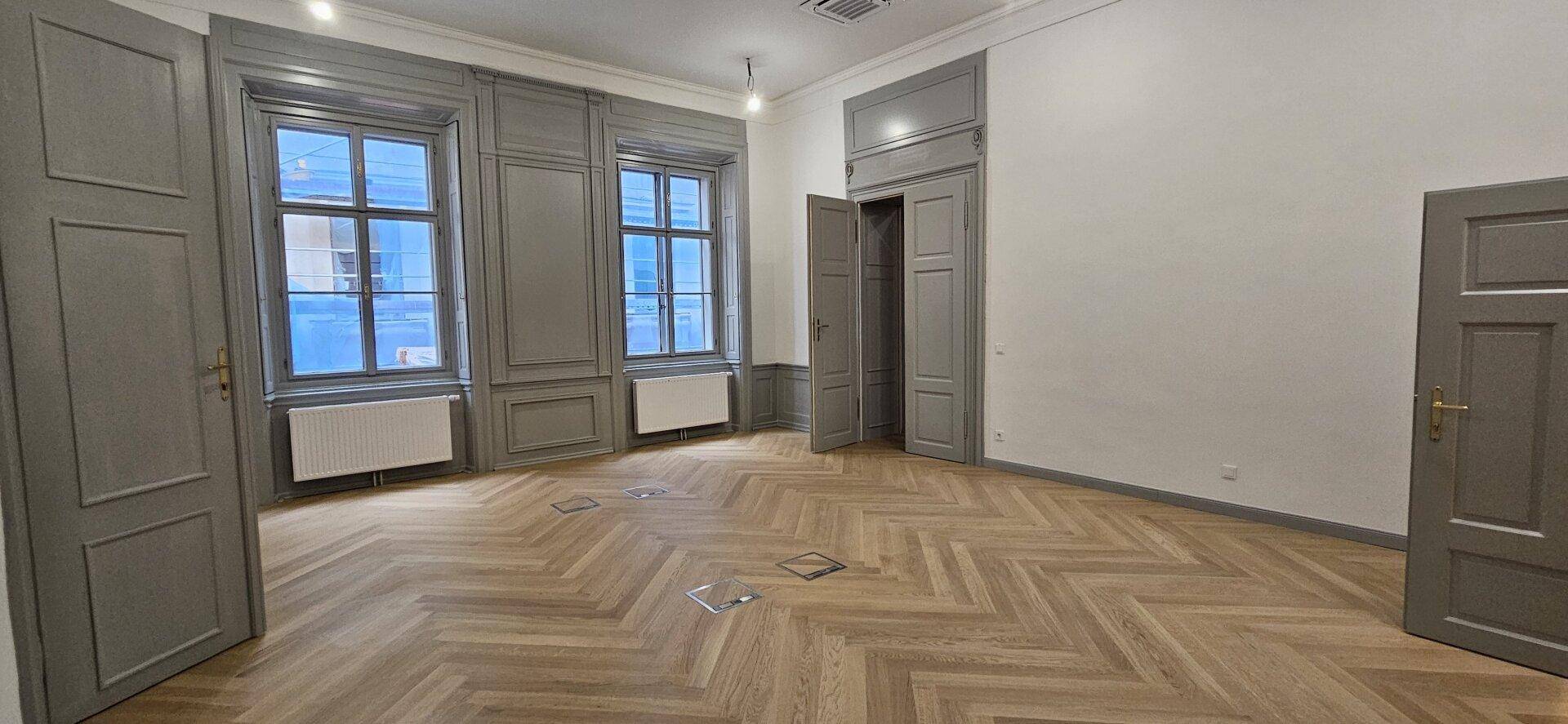 Heller Raum mit Fischgrätparkett, hohen Fenstern und dekorativen Wandpaneelen in Grau, sehr elegant.