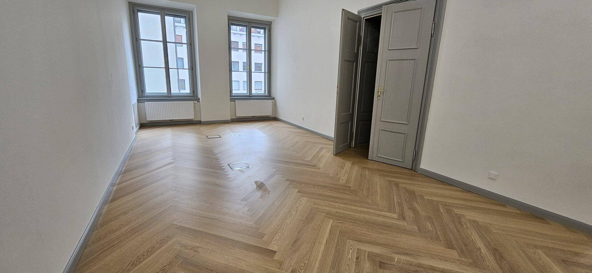 Geräumiger Raum mit Fischgrätparkett, hohen Fenstern und eleganten grauen Türen, vielseitig nutzbar.