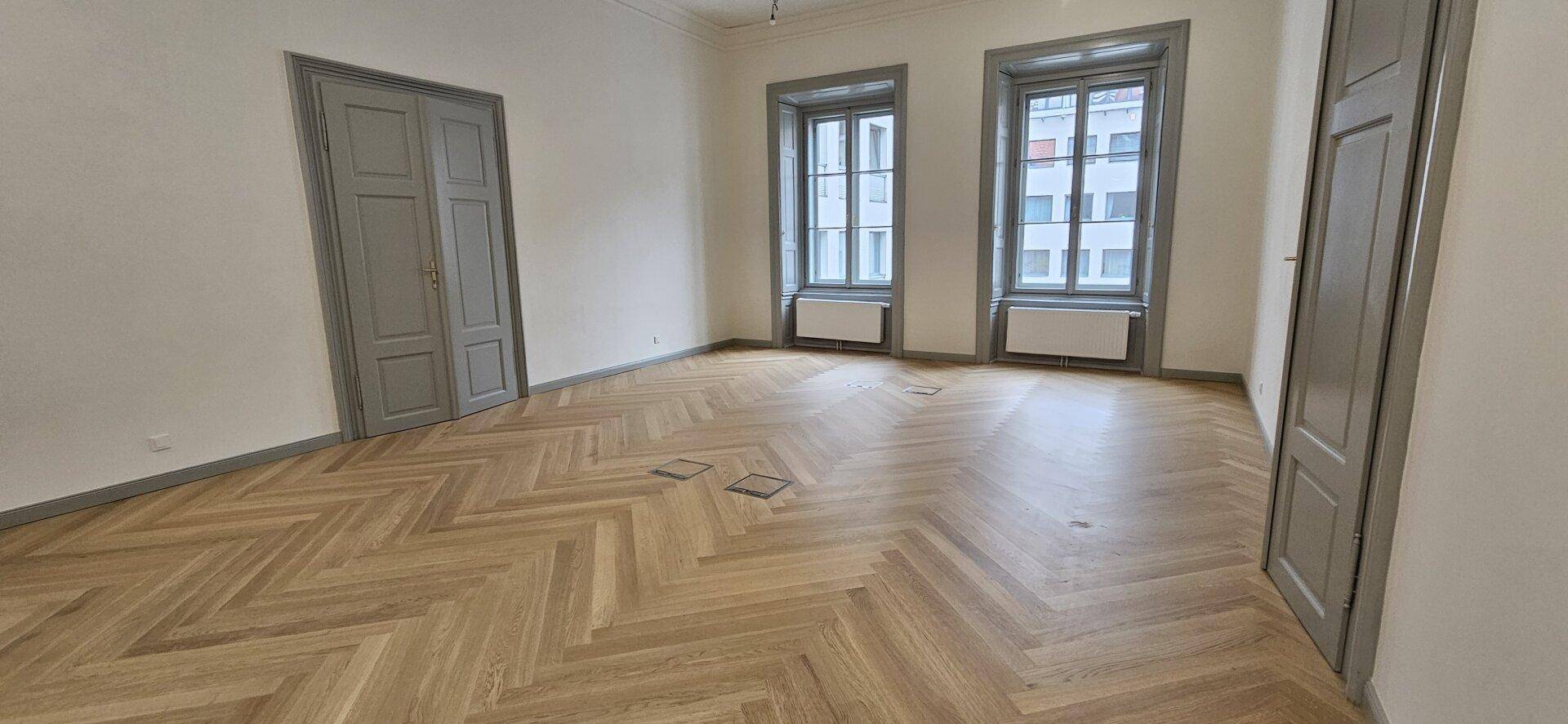 Geräumiger Raum mit Fischgrätparkett, hohen Fenstern und eleganten grauen Türen, vielseitig nutzbar.
