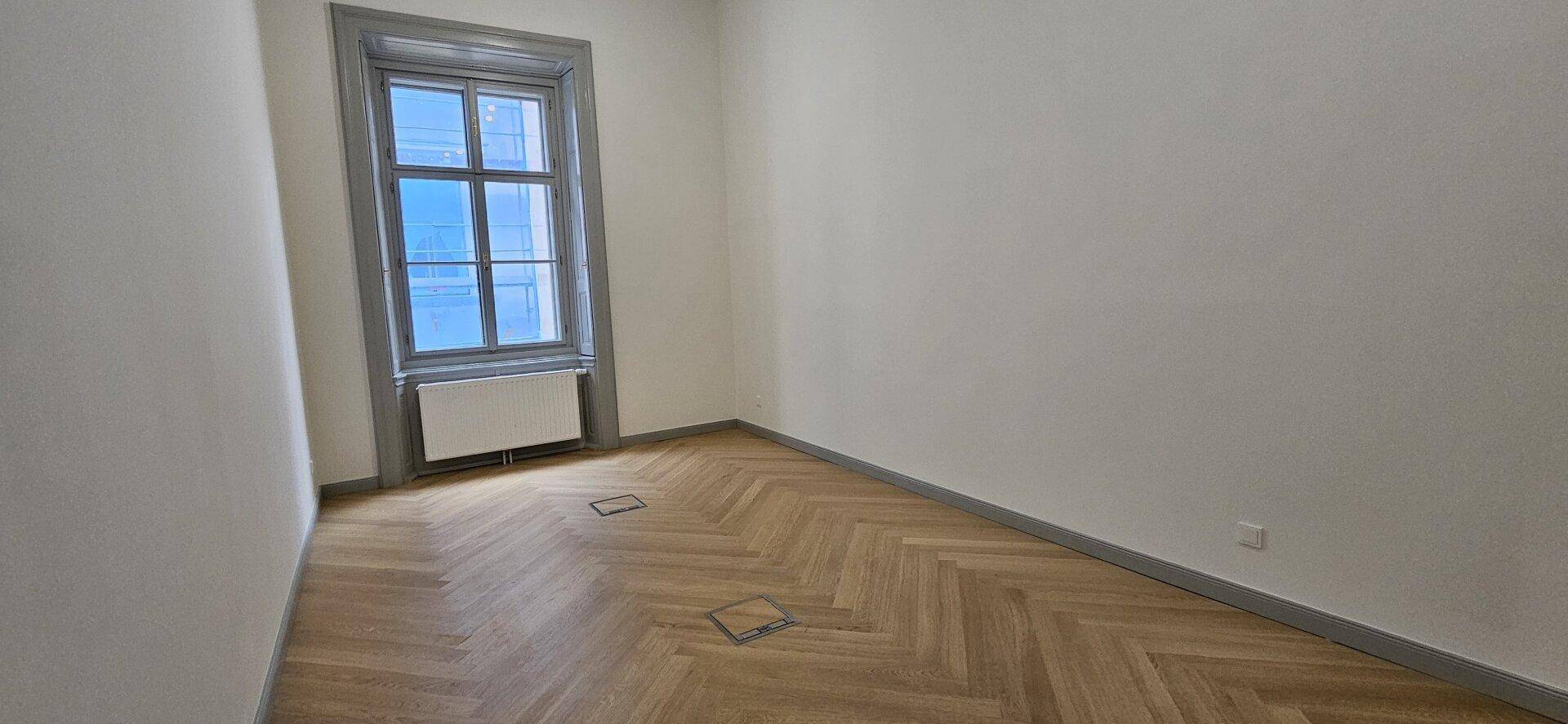 Heller Raum mit Fischgrätparkett, einem hohen Fenster und weiß gestrichenen Wänden, schlicht und elegant.