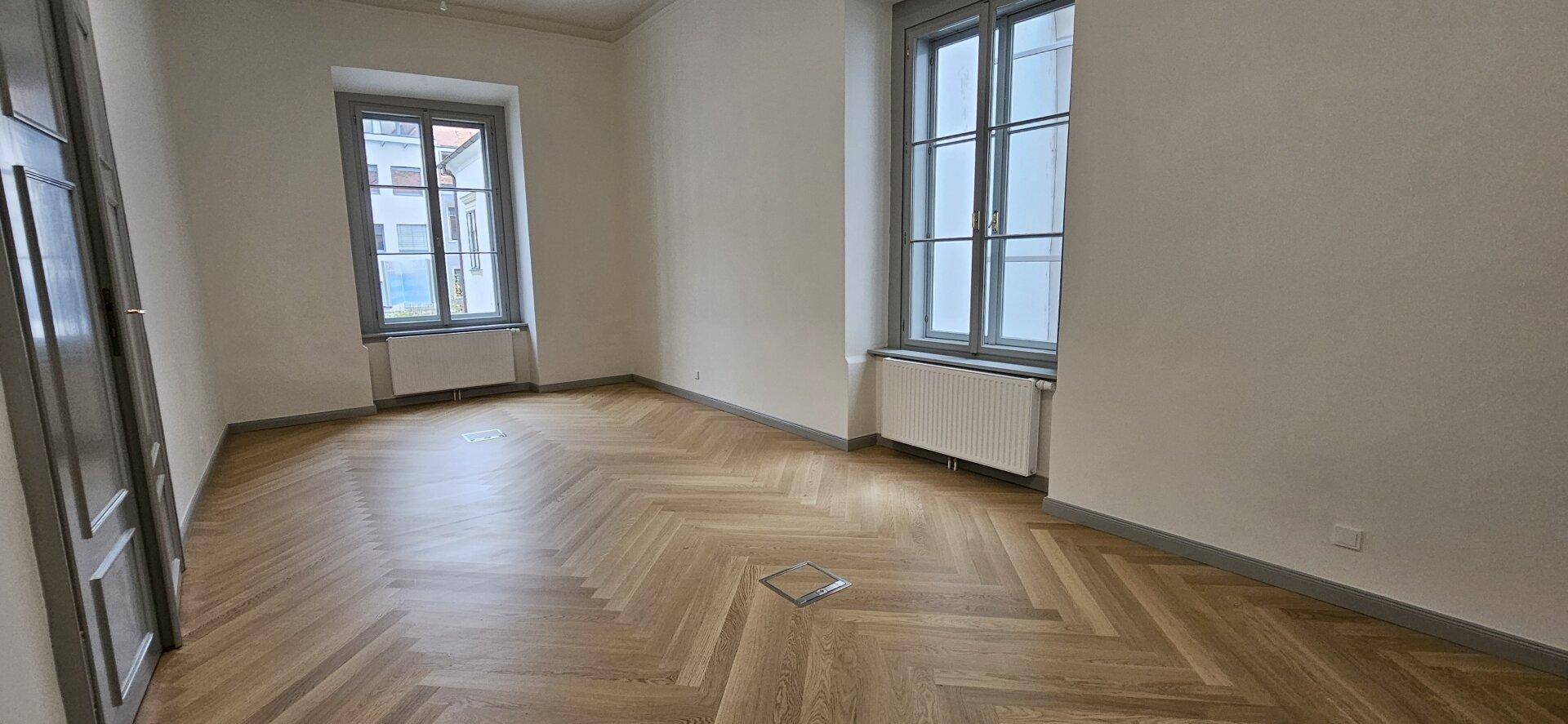 Heller Raum mit Fischgrätparkett, hohen Fenstern und weiß gestrichenen Wänden, bietet eine klare Ästhetik.