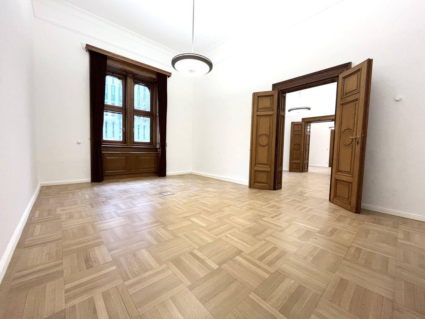 Heller Raum mit klassischem Parkettboden, hohen Decken und doppelten Holztüren, die weitere Räume verbinden.