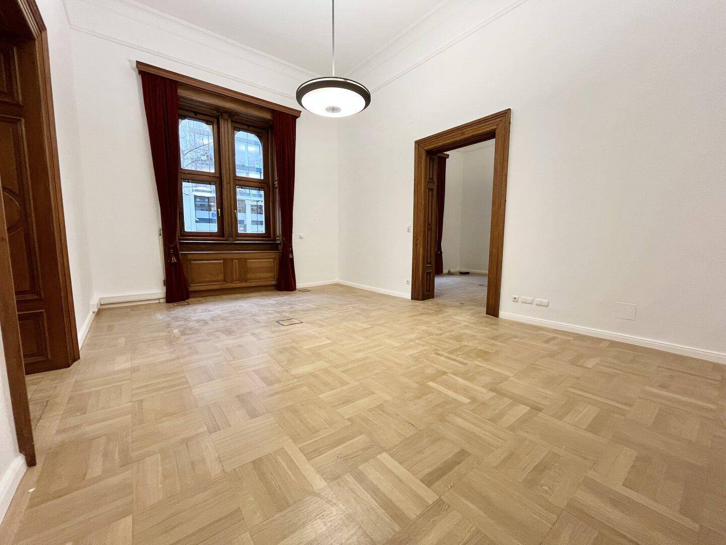 Großer, heller Raum mit elegantem Parkettboden und hohen Fenstern, die viel Tageslicht hereinlassen.