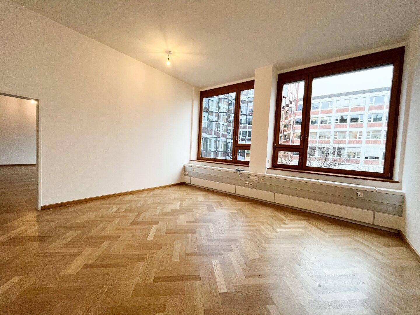 Leerer Raum mit hellem Fischgrätparkett und großen Fenstern, die viel Licht in den Raum bringen.