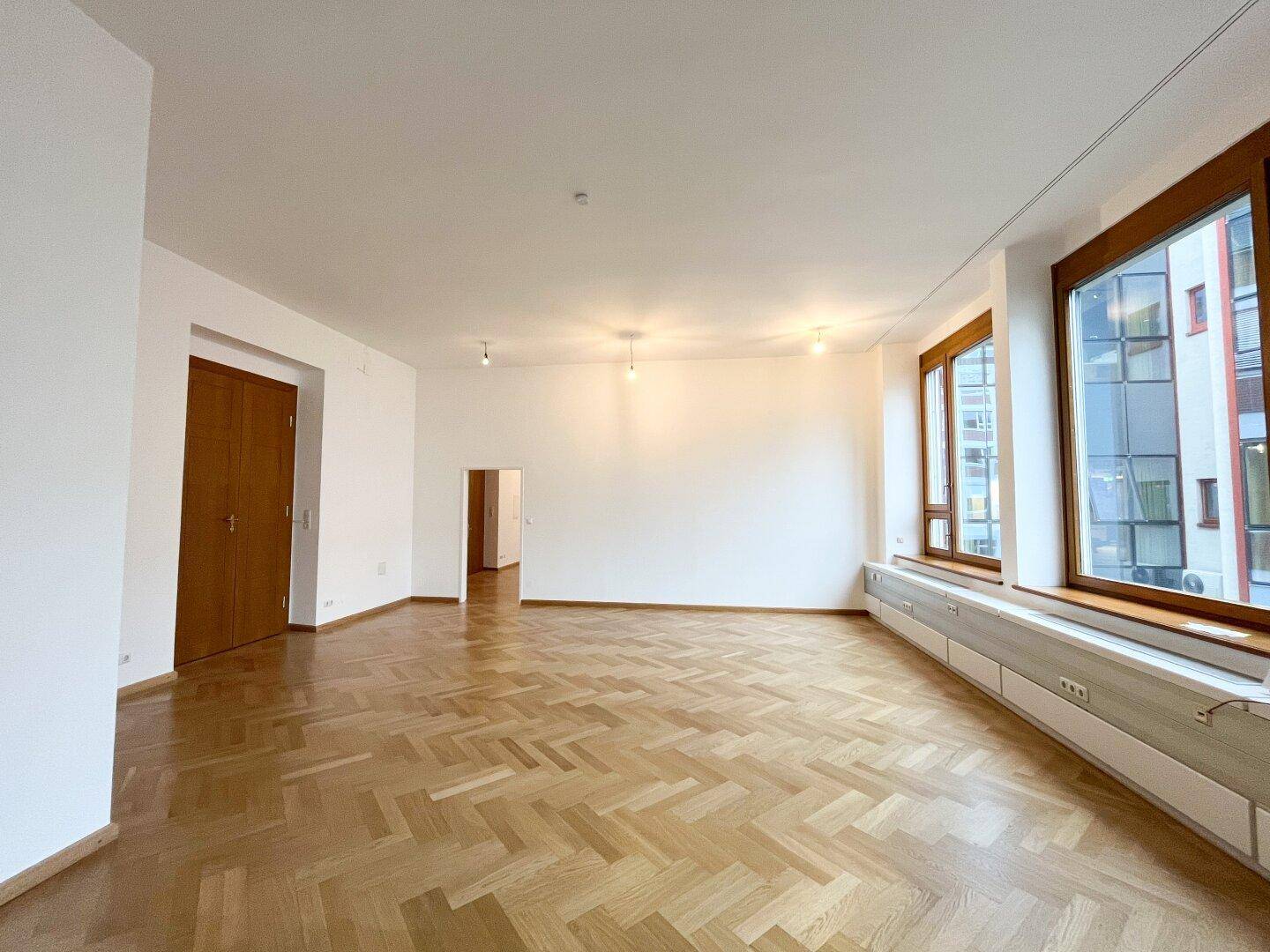 Großer, heller Raum mit Fischgrätparkett, hohen Decken und mehreren Fenstern.