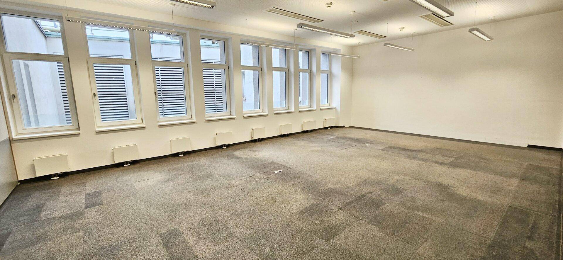 Großer, leerer Büroraum mit Teppichboden und einer Reihe von Fenstern, die Tageslicht hereinlassen.