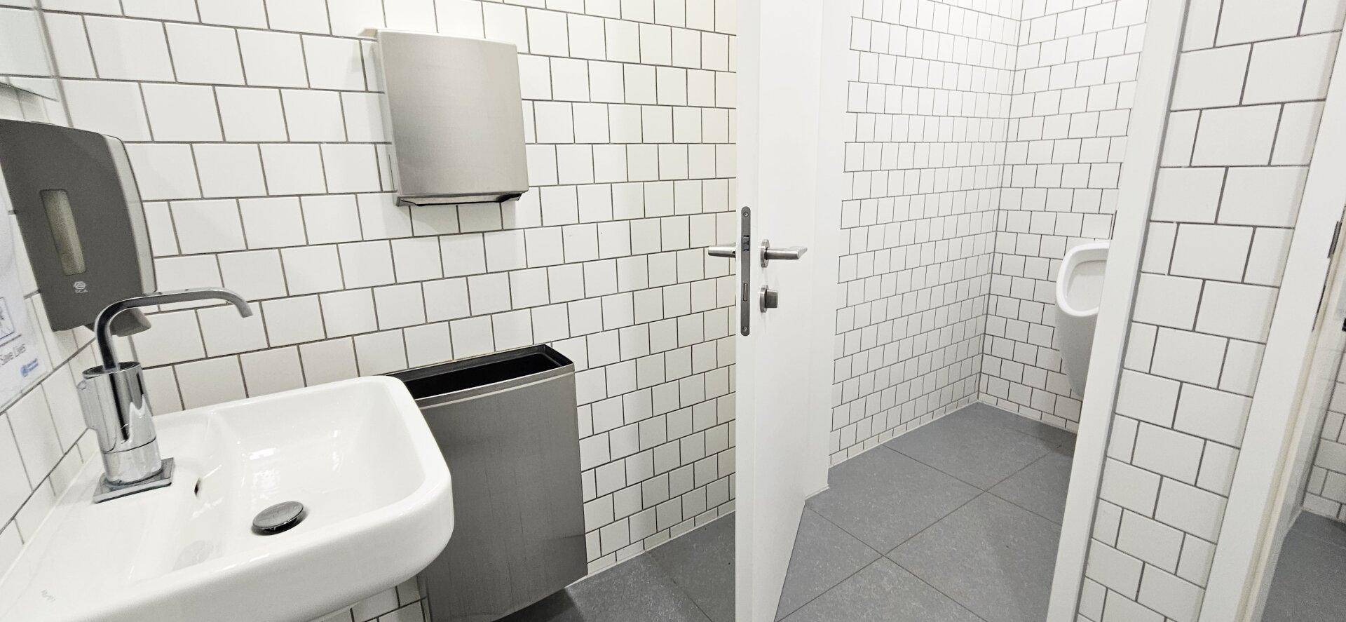 Modern gefliestes Badezimmer mit Waschbecken, Urinal und weißen Metrofliesen an den Wänden.