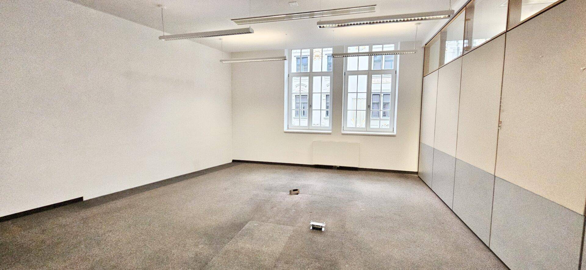 Leeres Büro mit Teppichboden, weißen Wänden und zwei großen Fenstern, die viel Licht spenden.