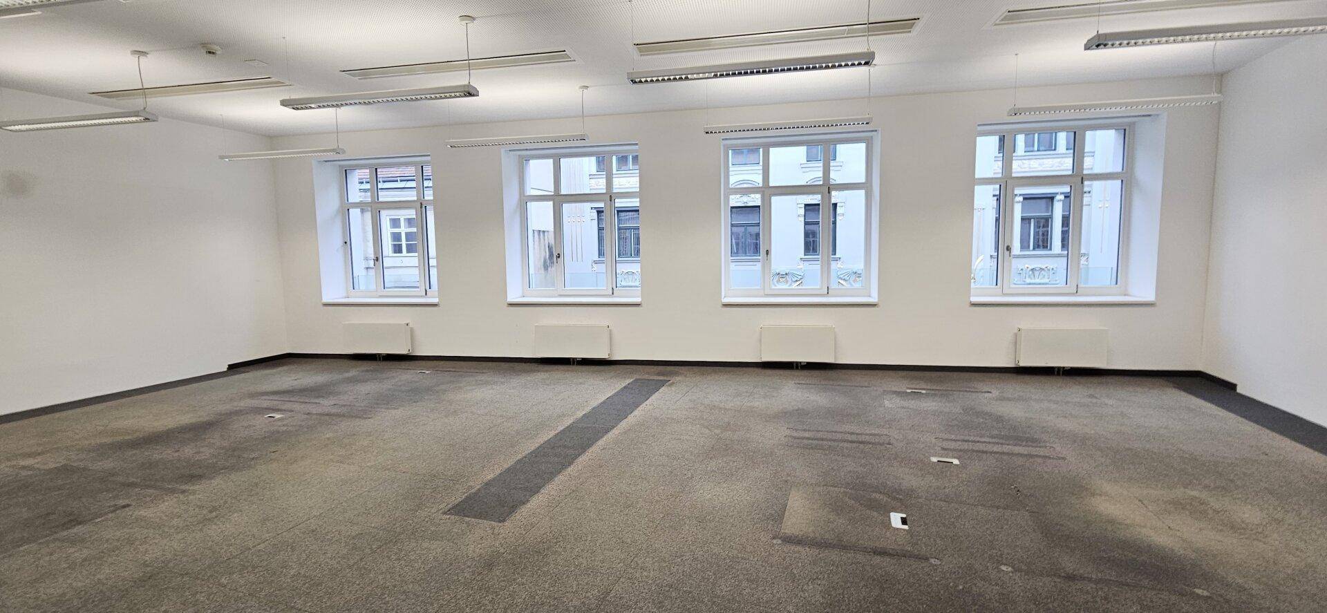 Heller Büroraum mit Teppichboden und einer Fensterfront, die den Raum gut belichtet.