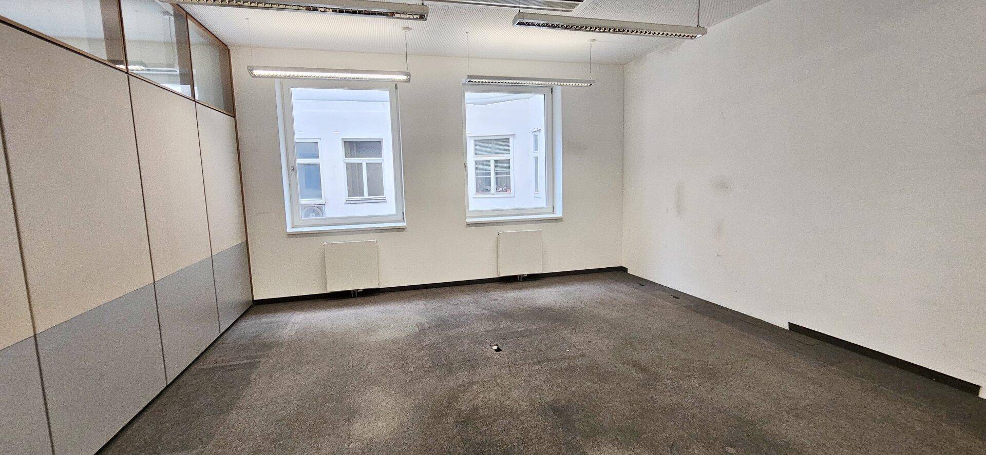Leeres Büro mit Teppichboden, weißen Wänden und zwei Fenstern, die den Raum mit Tageslicht versorgen.