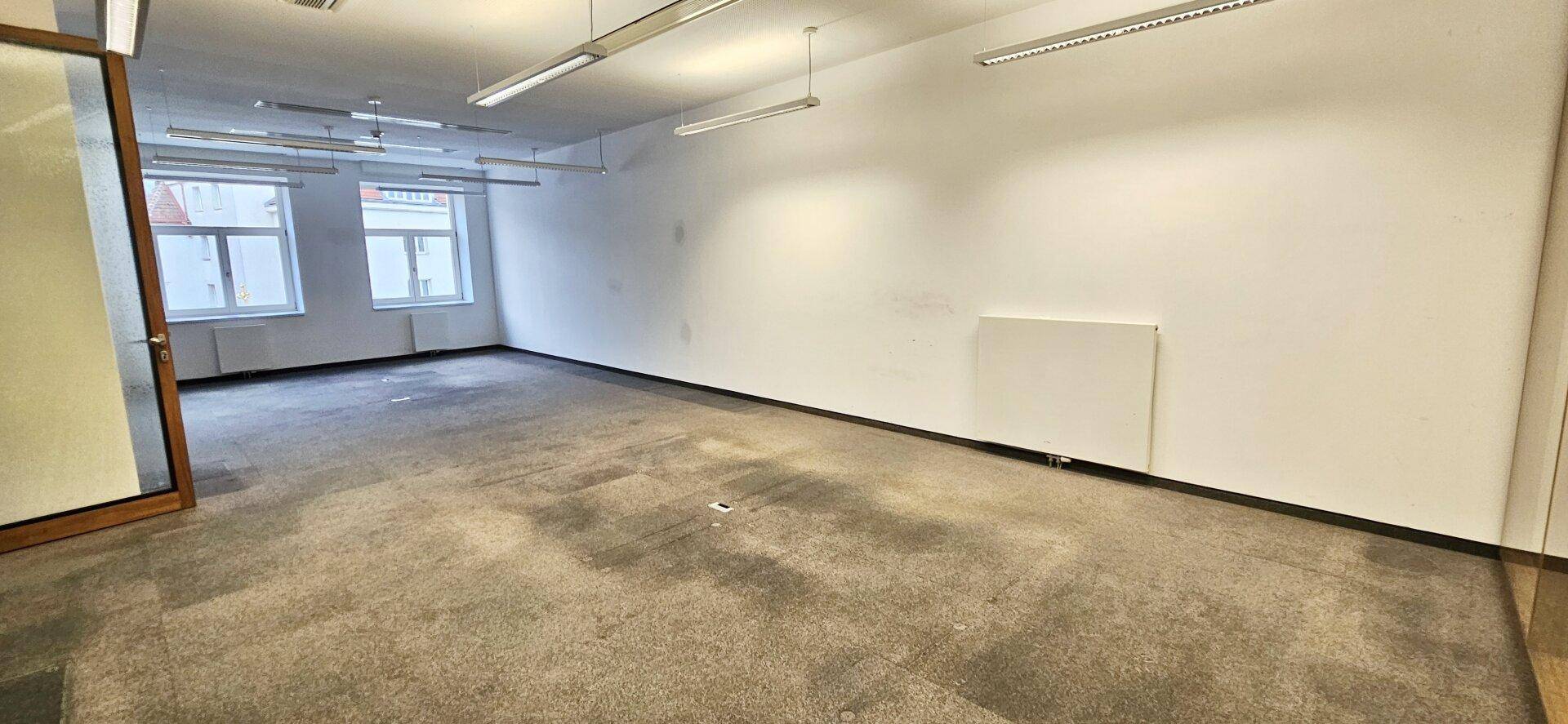 Großer, leerer Büroraum mit Teppichboden und einer langen Fensterreihe, die für viel Helligkeit sorgt.