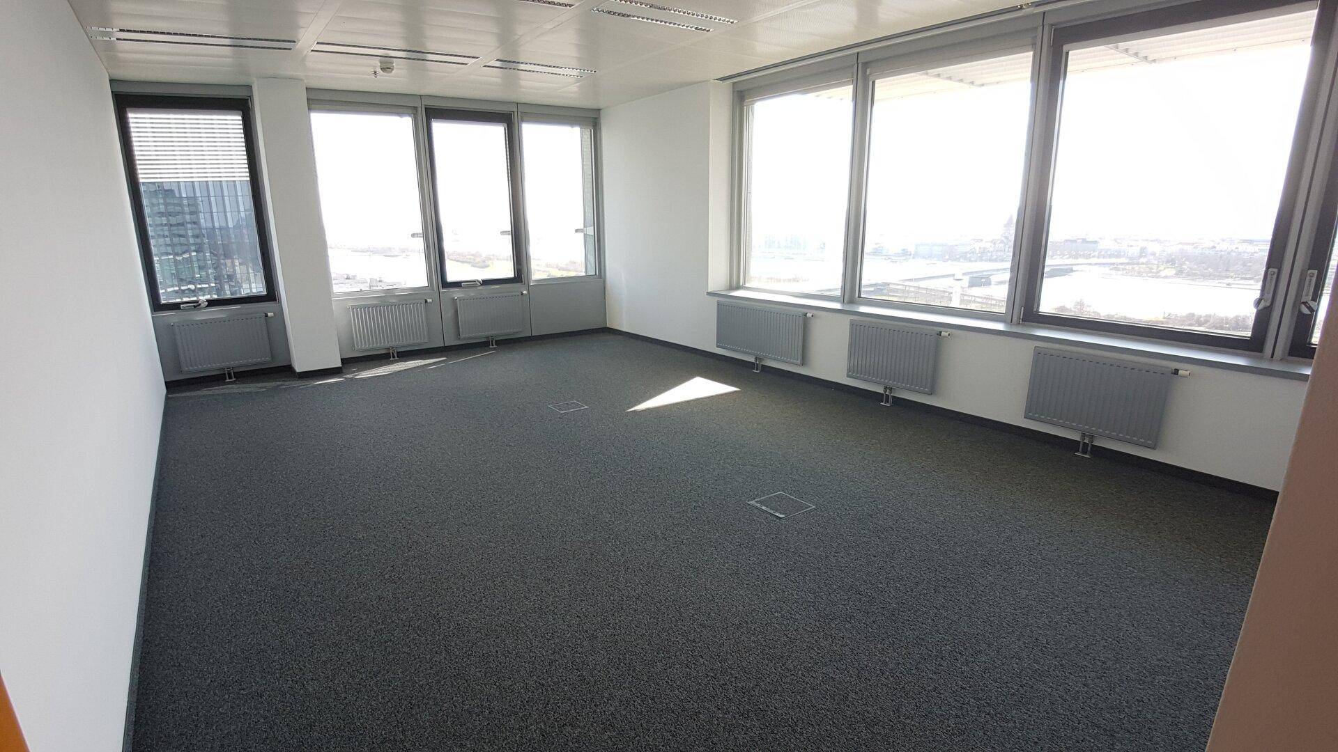 Leerer Büroraum mit großen Fenstern, Heizkörpern unter den Fenstern und dunklem Teppichboden.