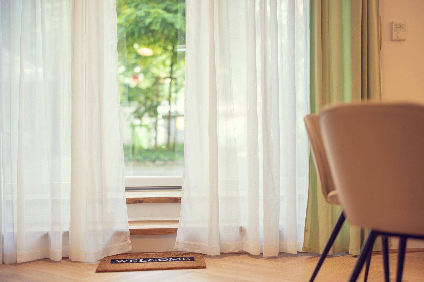 Große Fenster mit hellen Vorhängen und Blick ins Grüne, schaffen eine freundliche Atmosphäre.