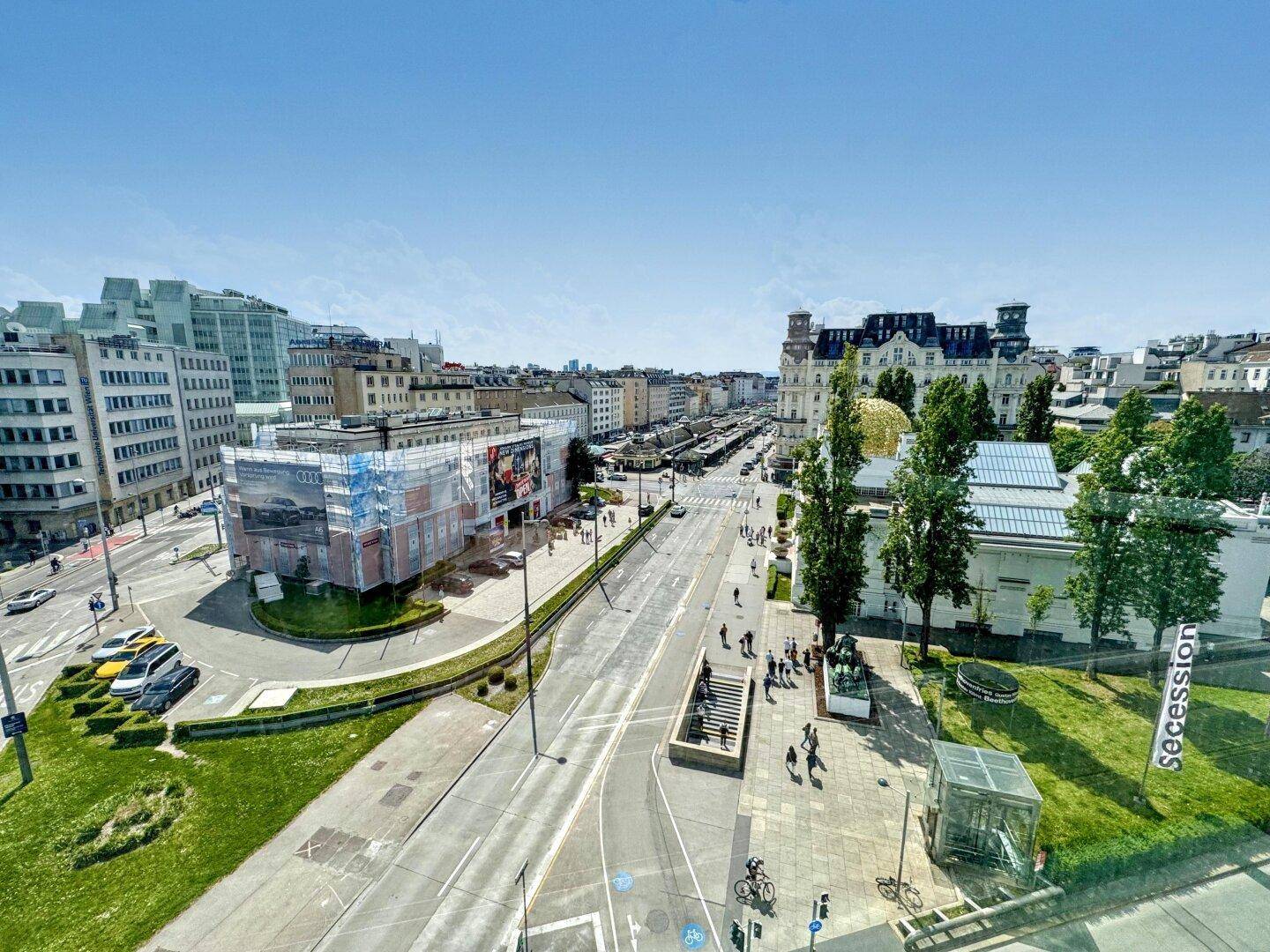 Weitläufiger Blick über eine belebte Stadtkreuzung mit Gebäuden und Grünflächen unter blauem Himmel.