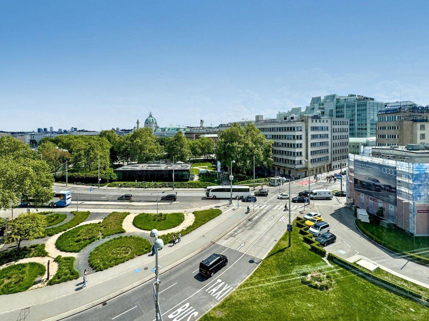 Panoramablick auf eine städtische Kreuzung mit Verkehr, Gebäuden und einem Park im Hintergrund.