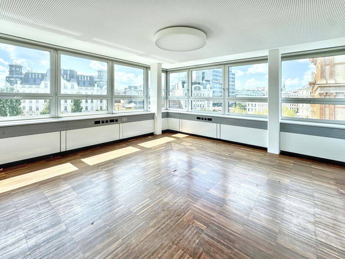 Leerer Büroraum mit Holzboden und großen Fenstern, die viel Tageslicht hereinlassen und Stadtblick bieten.