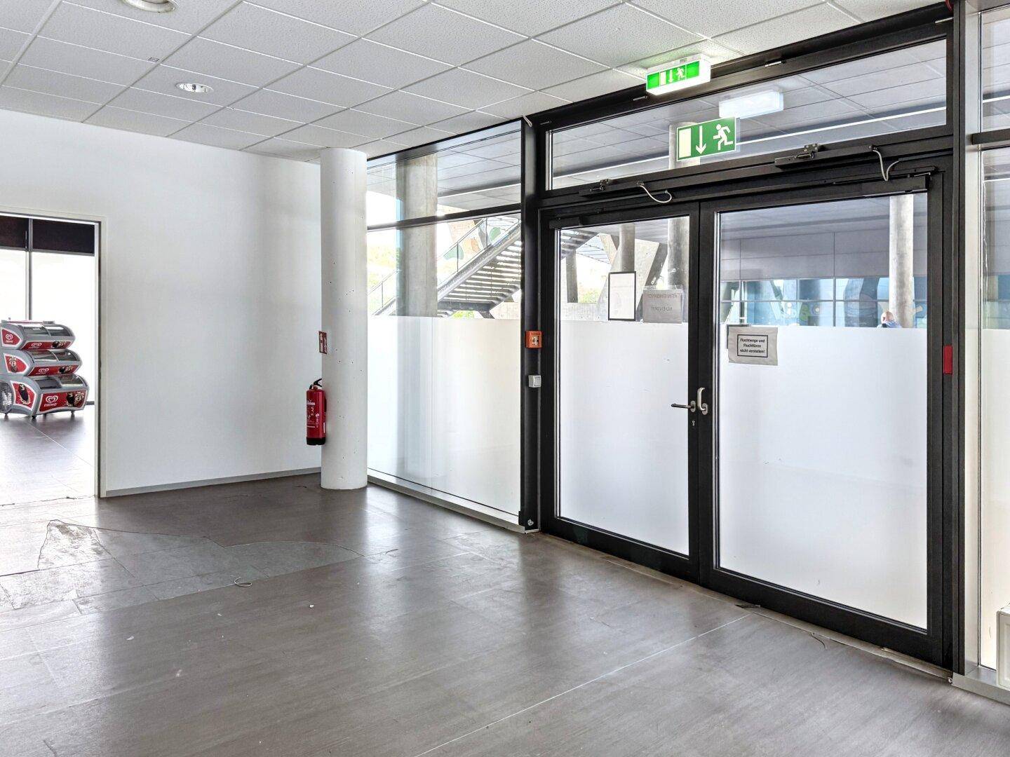 Eingangsbereich mit Glastüren, Brandschutz und einem Blick auf eine Treppe im Hintergrund.