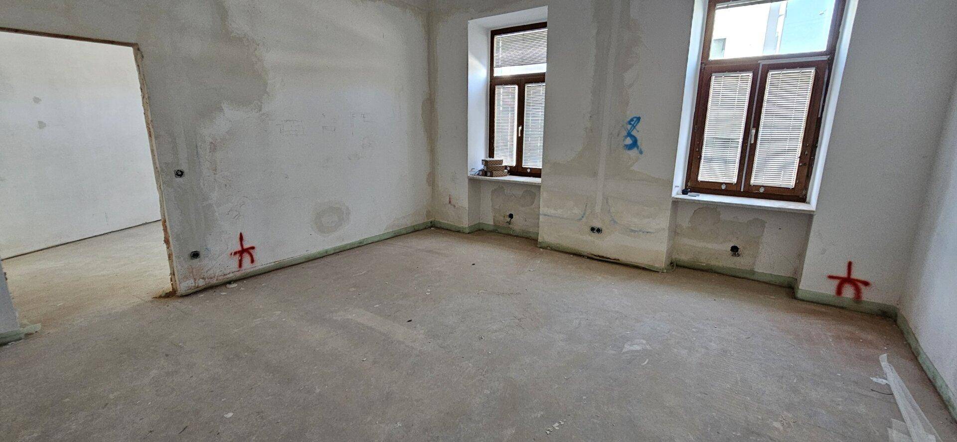 Leerer Raum mit unverputzten Wänden, Betonboden und zwei Fenstern mit Holzrahmen, bereit für den Ausbau.