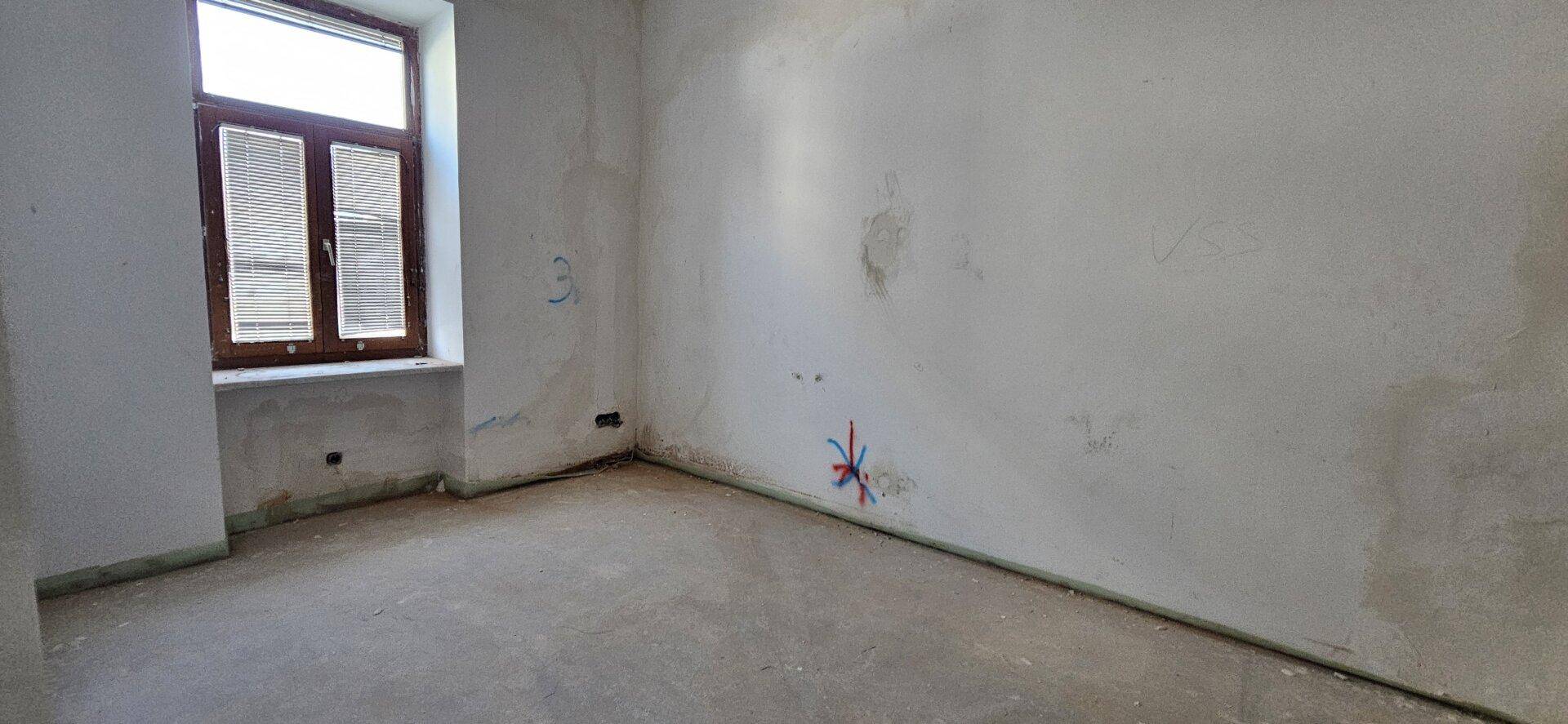Leerer Raum mit unverputzten Wänden, Betonboden und einem Fenster mit Holzrahmen, ideal für individuelle Gestaltung.