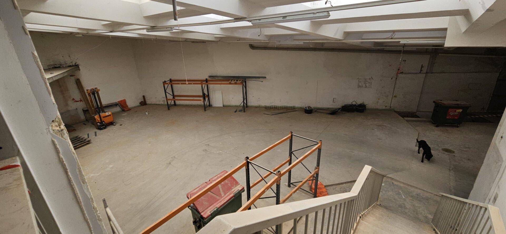 Blick von einer Treppe in den weitläufigen Industrieraum mit Betonboden und hohen Decken, noch im Rohbau.
