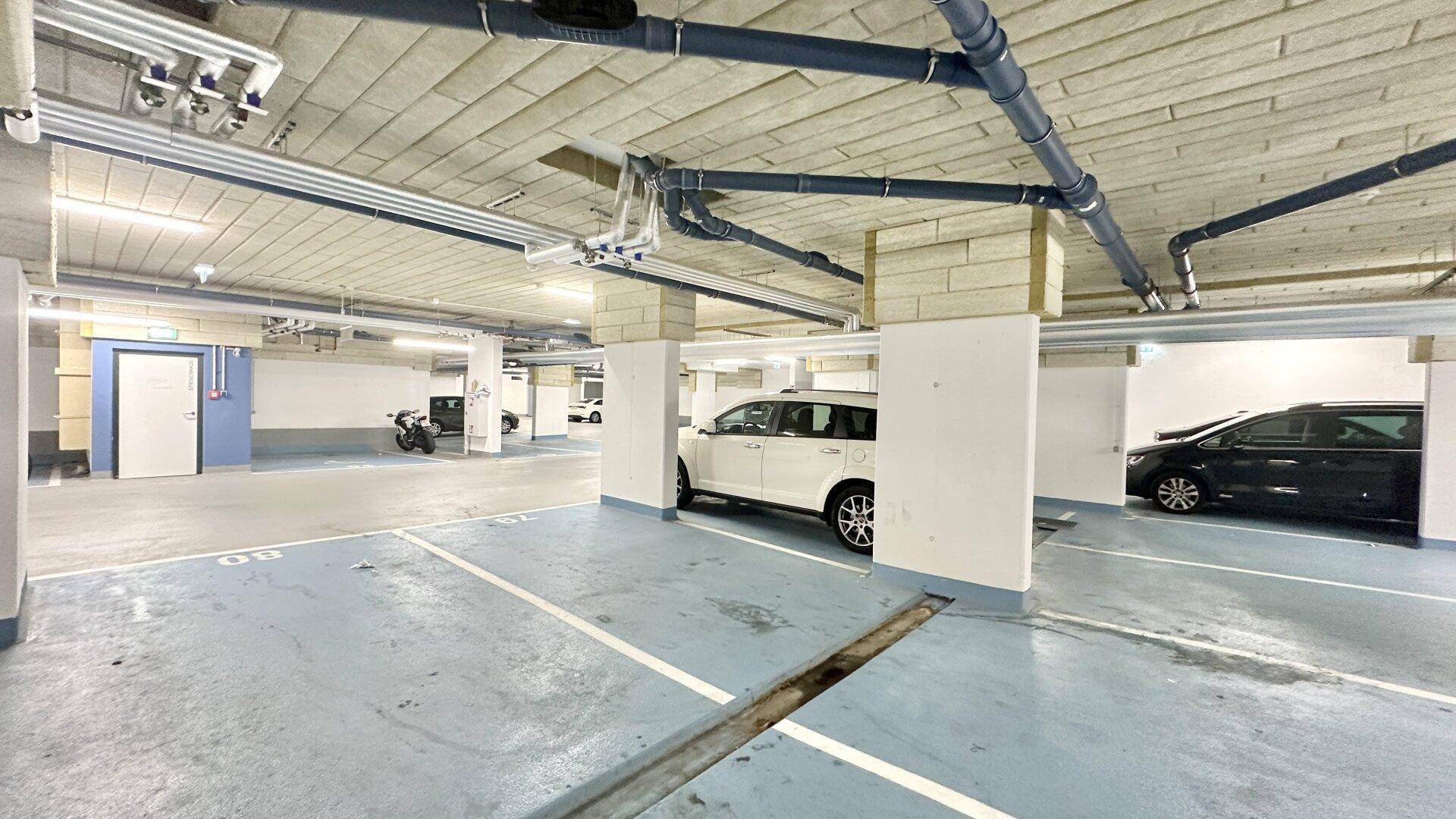 Geräumige Tiefgarage mit mehreren Fahrzeugen, sichtbaren Rohren an der Decke und Säulen.