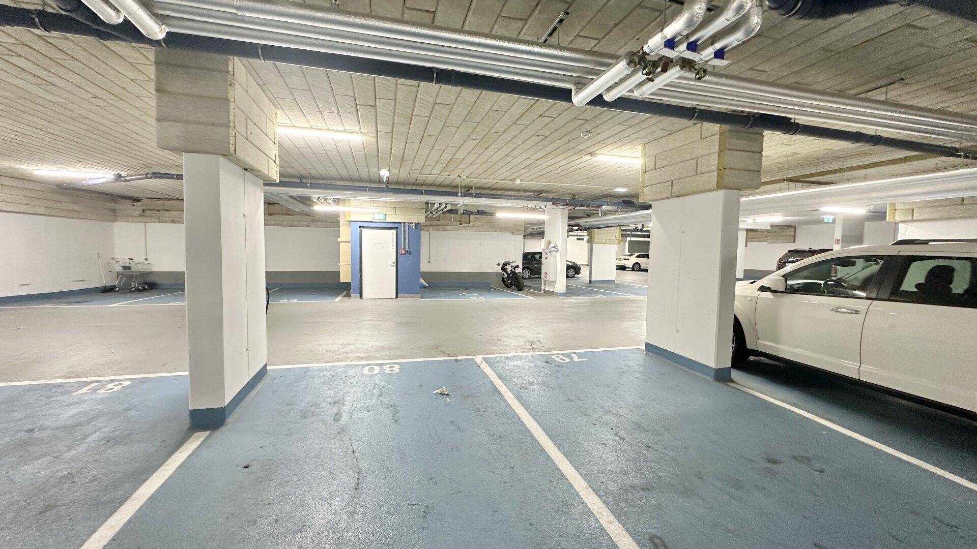 Helle Tiefgarage mit blauen Bodenmarkierungen, Säulen und sichtbaren Installationen an der Decke.