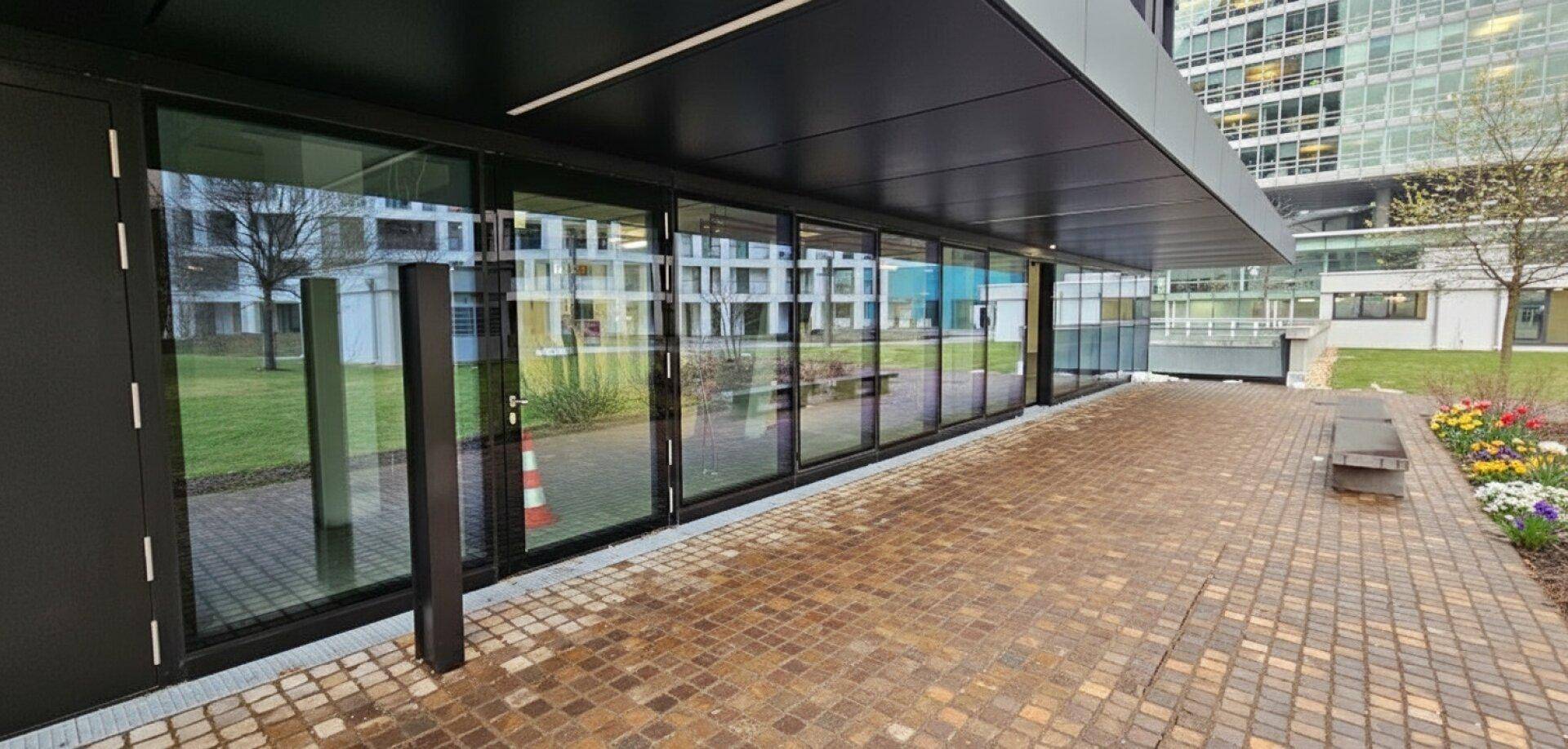 Moderne Gebäudefassade mit großen Glasfenstern und einem gepflasterten Eingangsbereich im Freien.